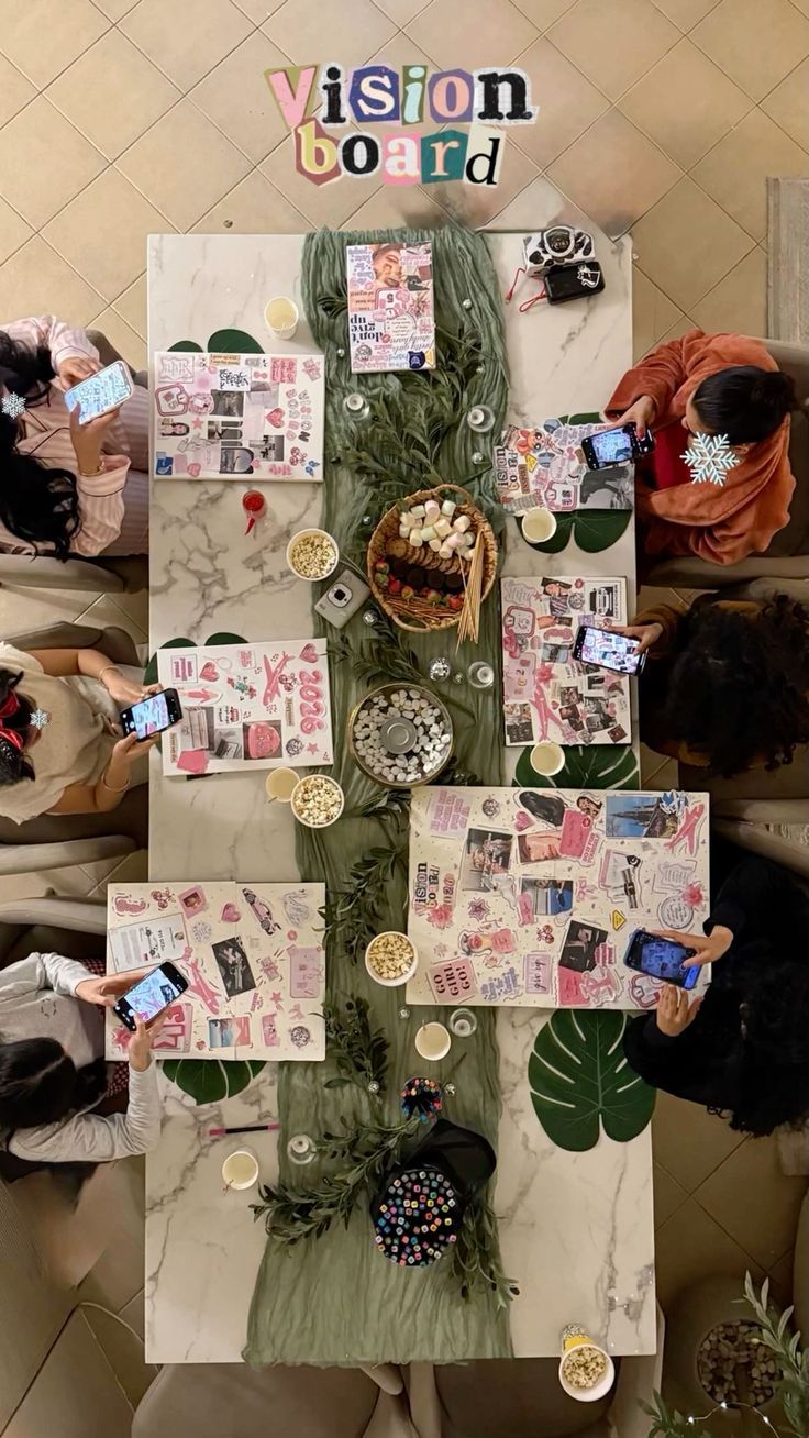 Overhead view of a vision board party with collages, snacks, and phones on a marble table.