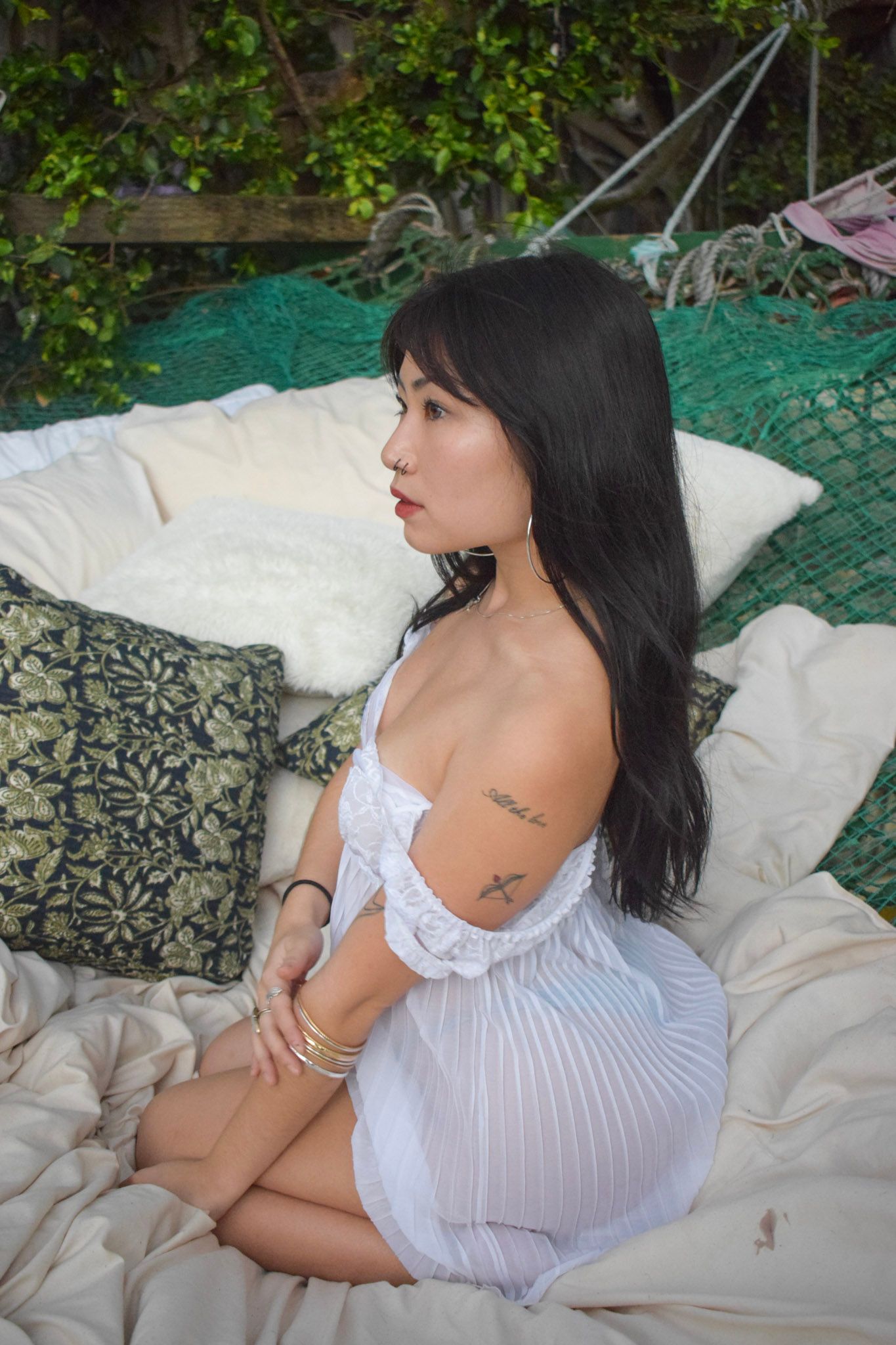 Figure in white dress reclining on outdoor cushions with greenery in background.