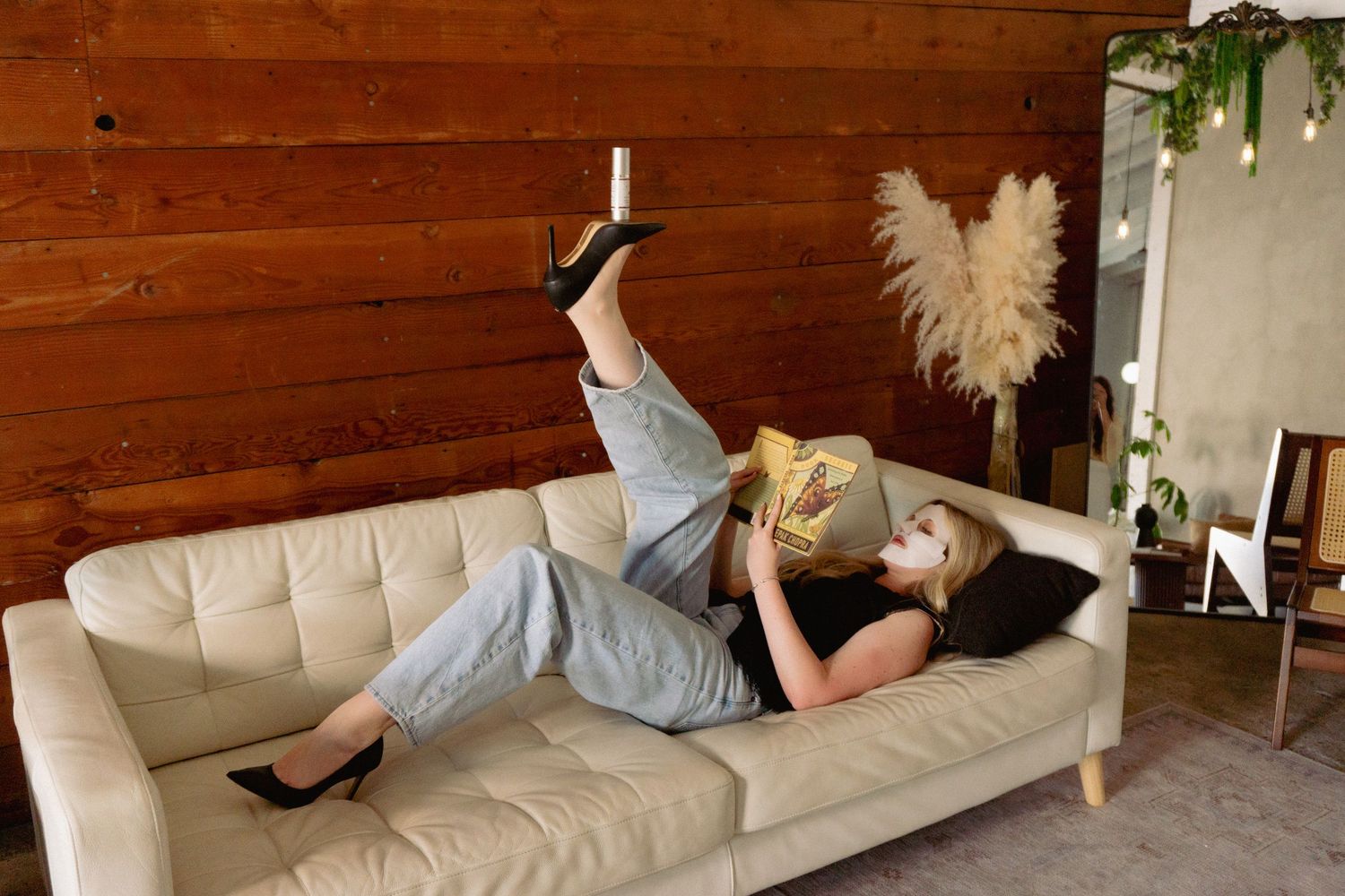 Woman in gray jumpsuit and heels lying on beige sofa reading a book against rustic wood panel wall.
