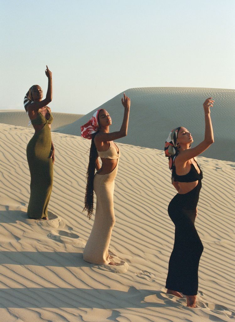 Three women in elegant dresses pose with raised arms on sand dunes at sunset.