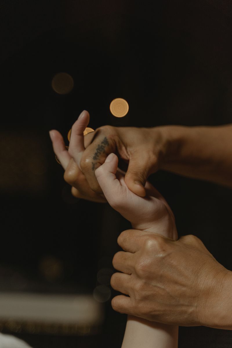Close-up of two hands with a tattoo gently holding another persons hand against a dark bokeh background.