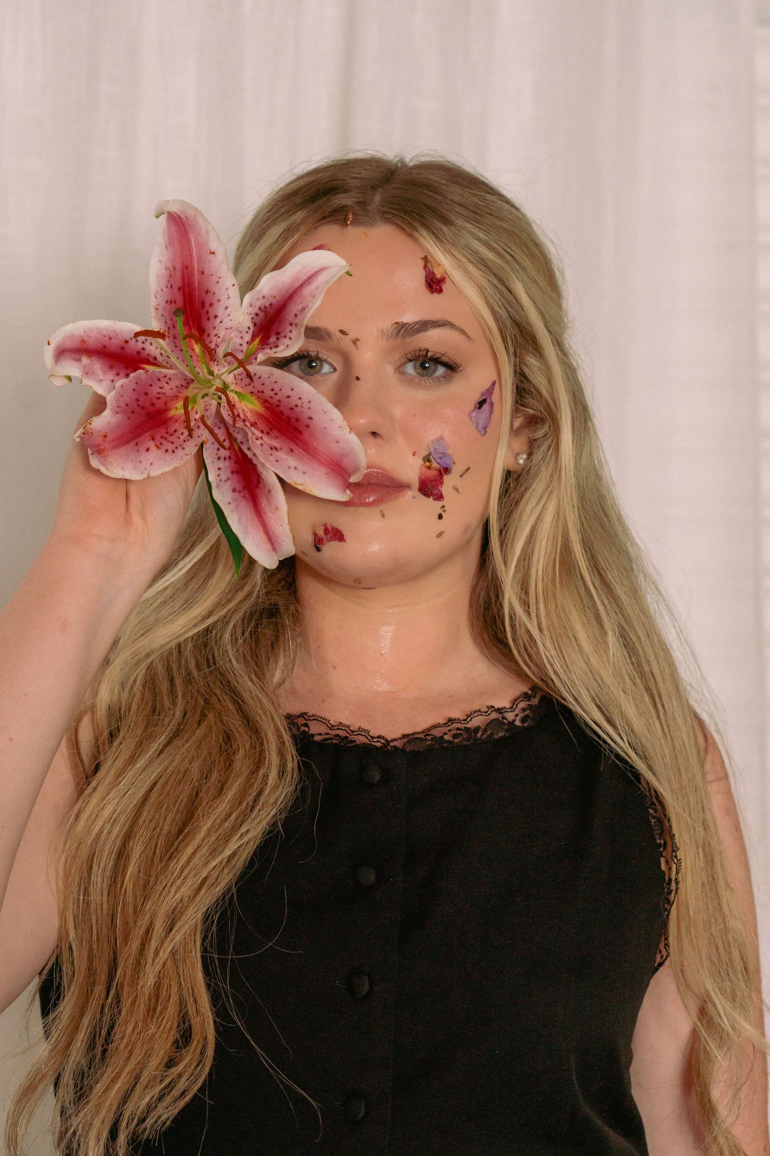 Blonde woman holding a pink stargazer lily over her eye with colorful floral petals on her face, wearing a black top.