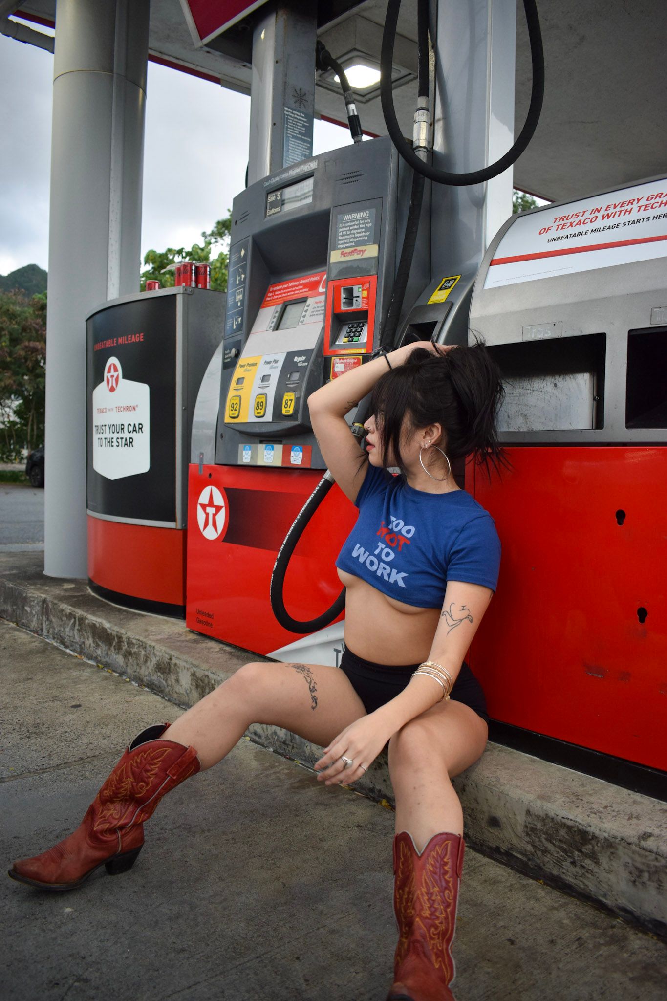 Woman in cowboy boots posing playfully at a gas station with red fuel pumps.