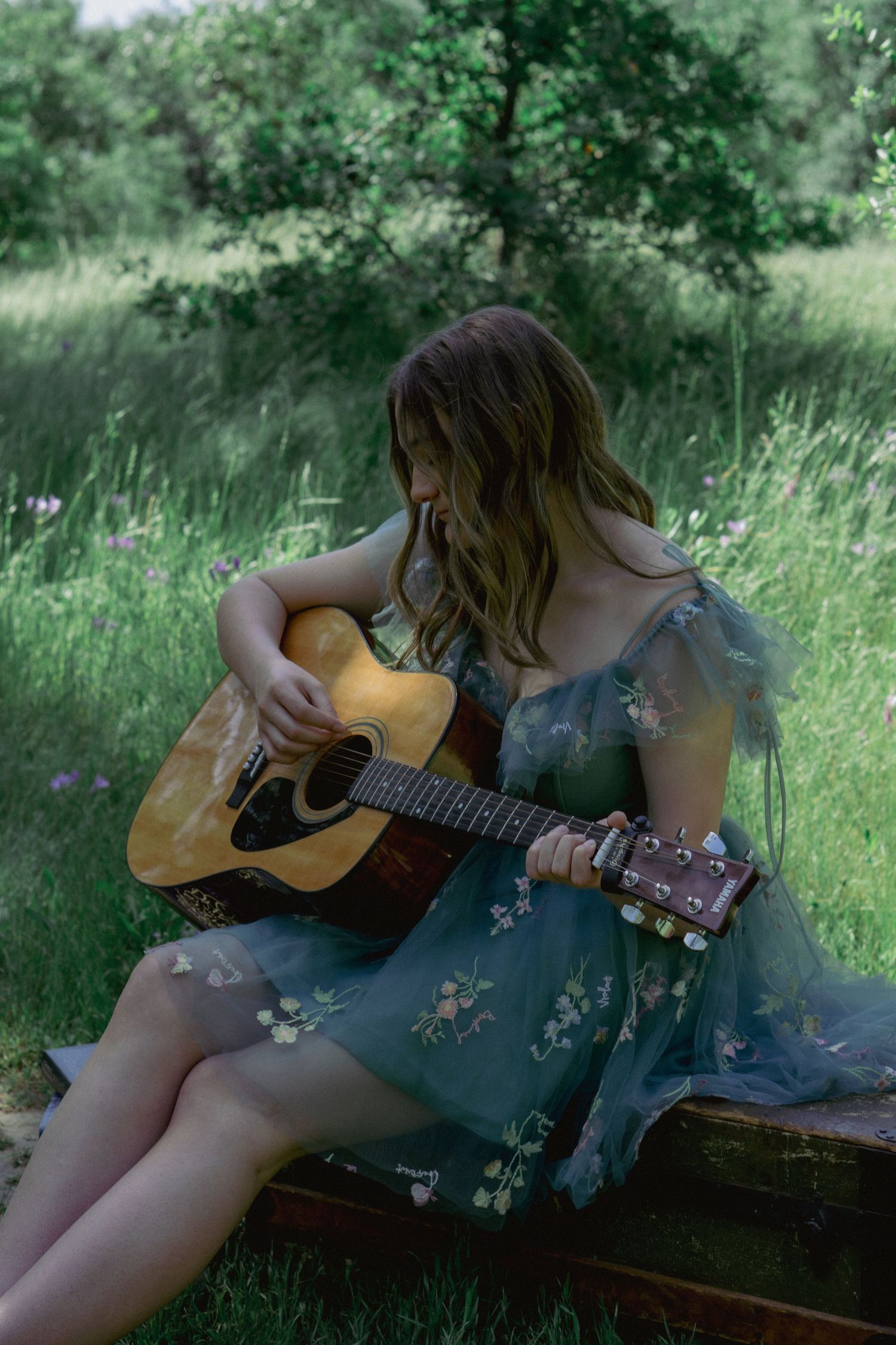 A figure in a floral turquoise dress plays acoustic guitar while sitting outdoors in tall grass.