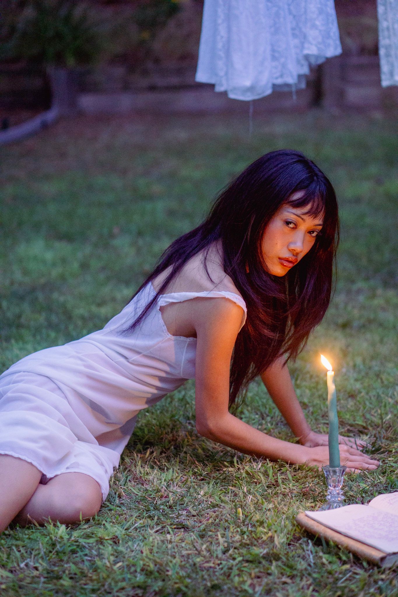 Person in white dress lying on grass near lit candle at dusk.