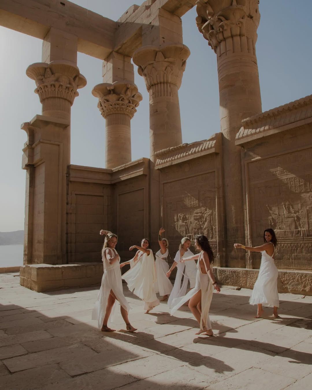 A group in white dresses dances and poses between ancient Egyptian temple columns.
