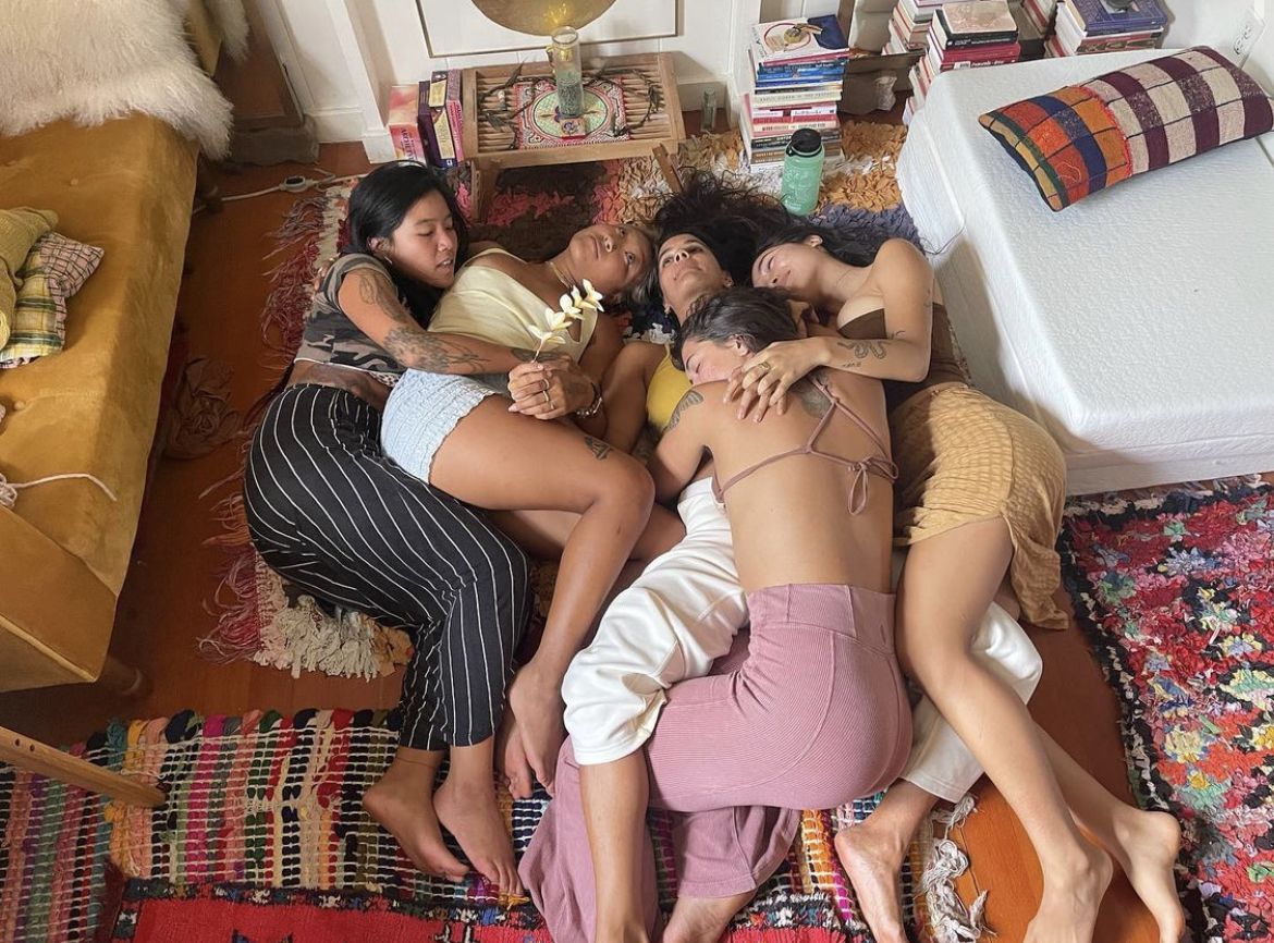 Four young women lying together on colorful rugs on a floor, relaxing and embracing in a cozy bohemian room.