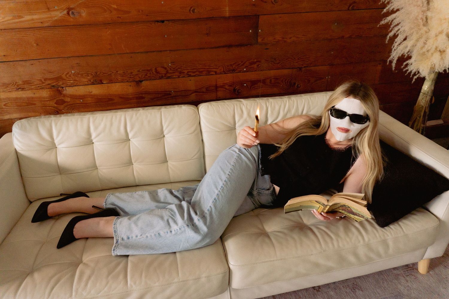 Young woman in sunglasses relaxing on cream sofa, reading a book with long blonde hair, wearing black top and gray pants.