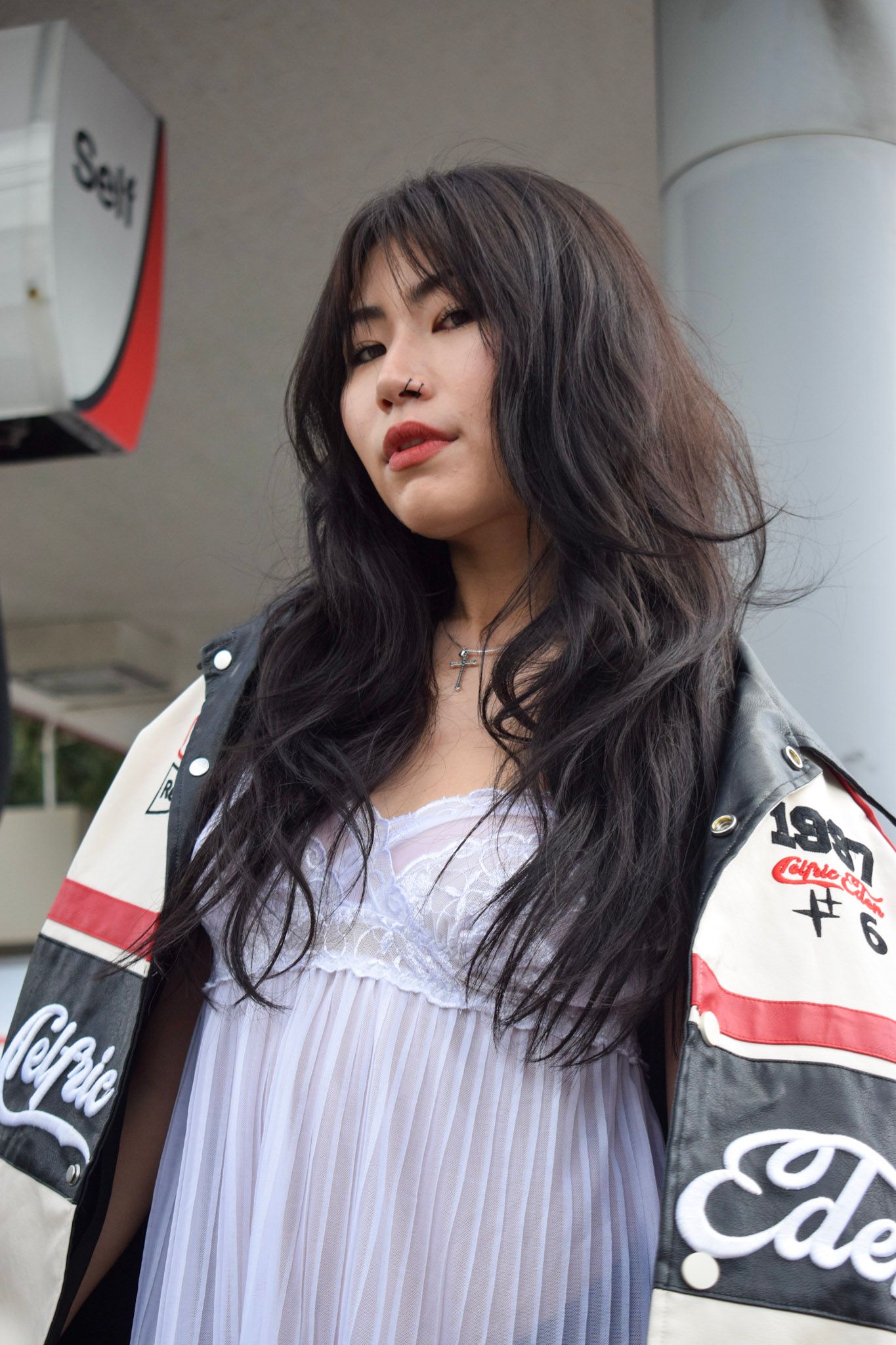 Portrait in white dress and leather jacket with long dark hair against urban backdrop.