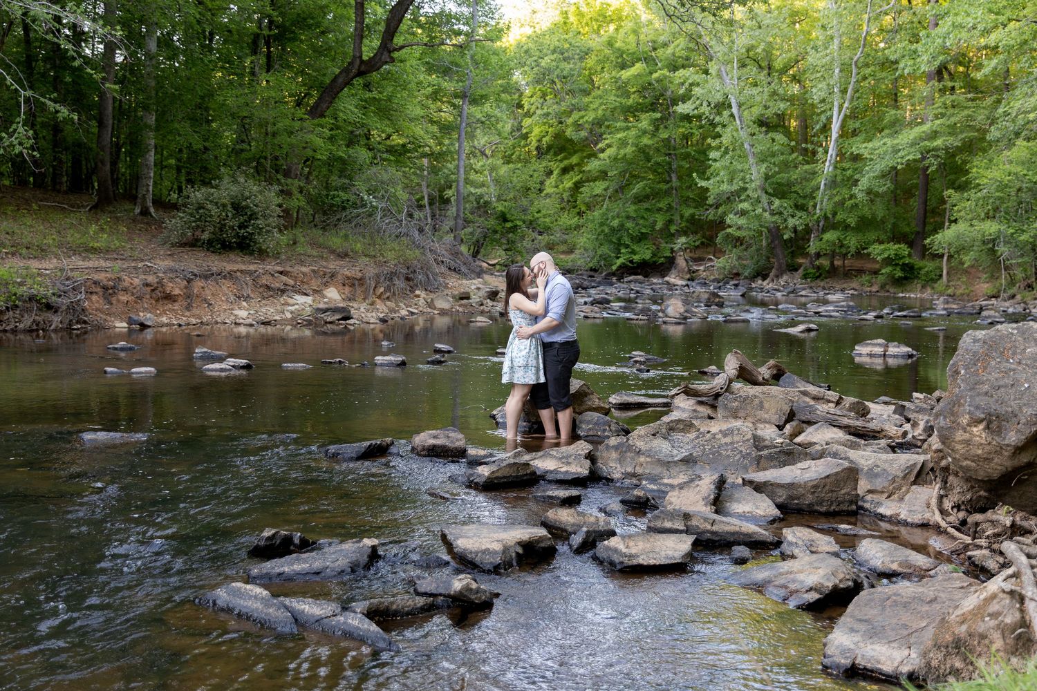Couple hugging on rocky stones in a shallow forest creek surrounded by lush trees.