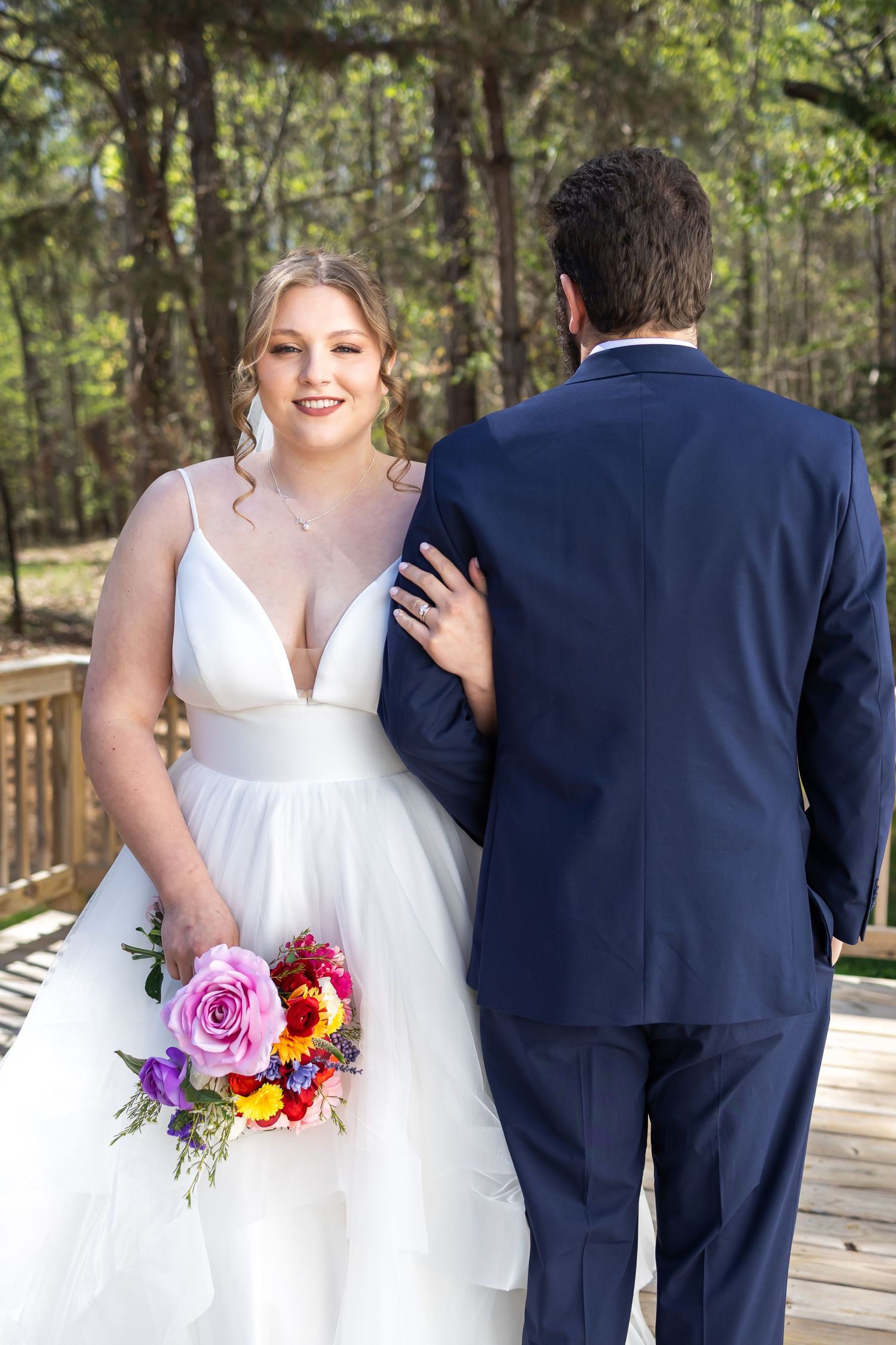 Blonde bride in white gown holding colorful bouquet standing with groom in navy suit on wooden deck.