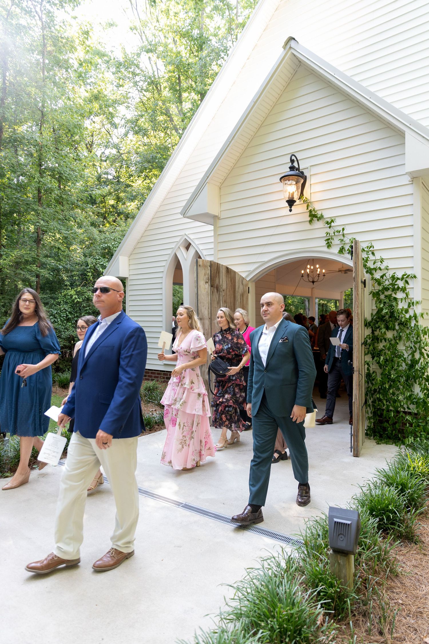 Wedding guests in formal attire walk outside a white wooden chapel with an arched doorway, surrounded by green trees and landscaped plants.