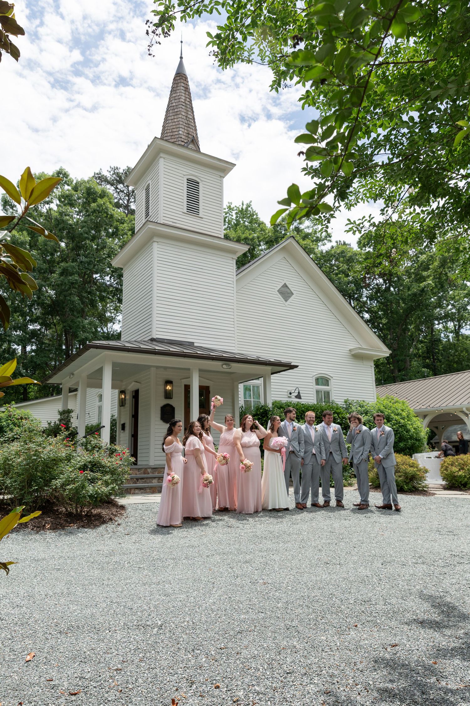 Wedding party poses in pink bridesmaid dresses and gray suits before white chapel with steeple.