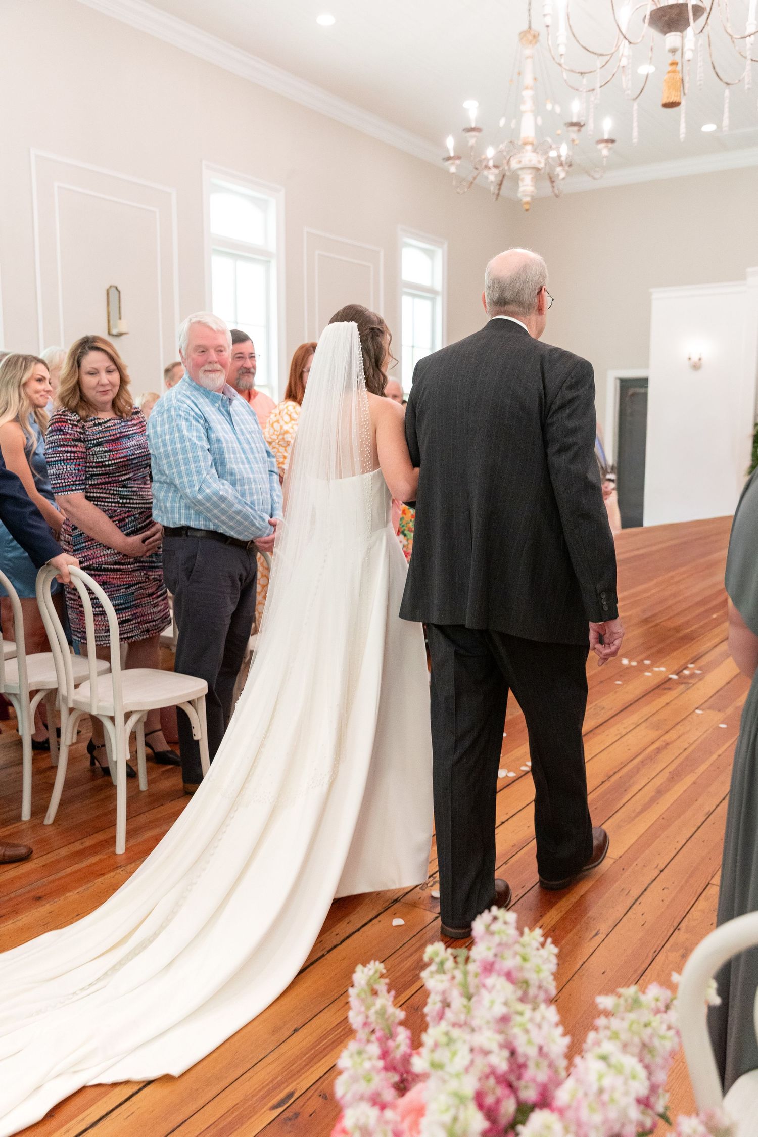 Bride in a long veil and gown walks down the wooden aisle of a chandelier-lit venue as seated wedding guests look on.