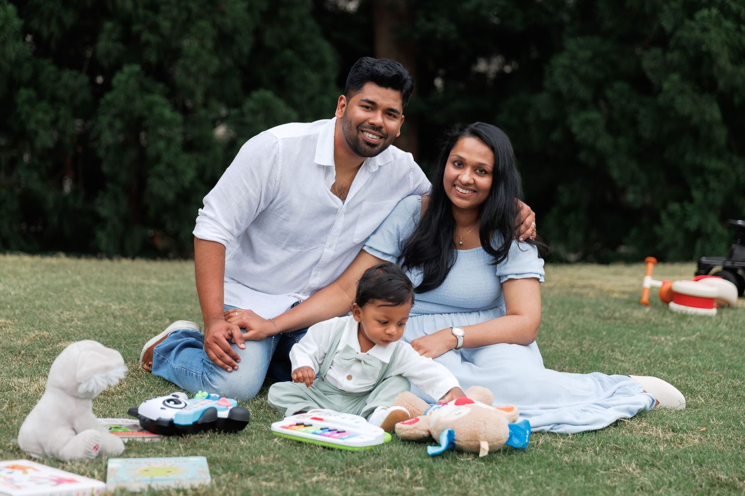 Smiling couple sitting on a grassy lawn with their baby, surrounded by plush toys and books, in a sunny park with green trees behind.