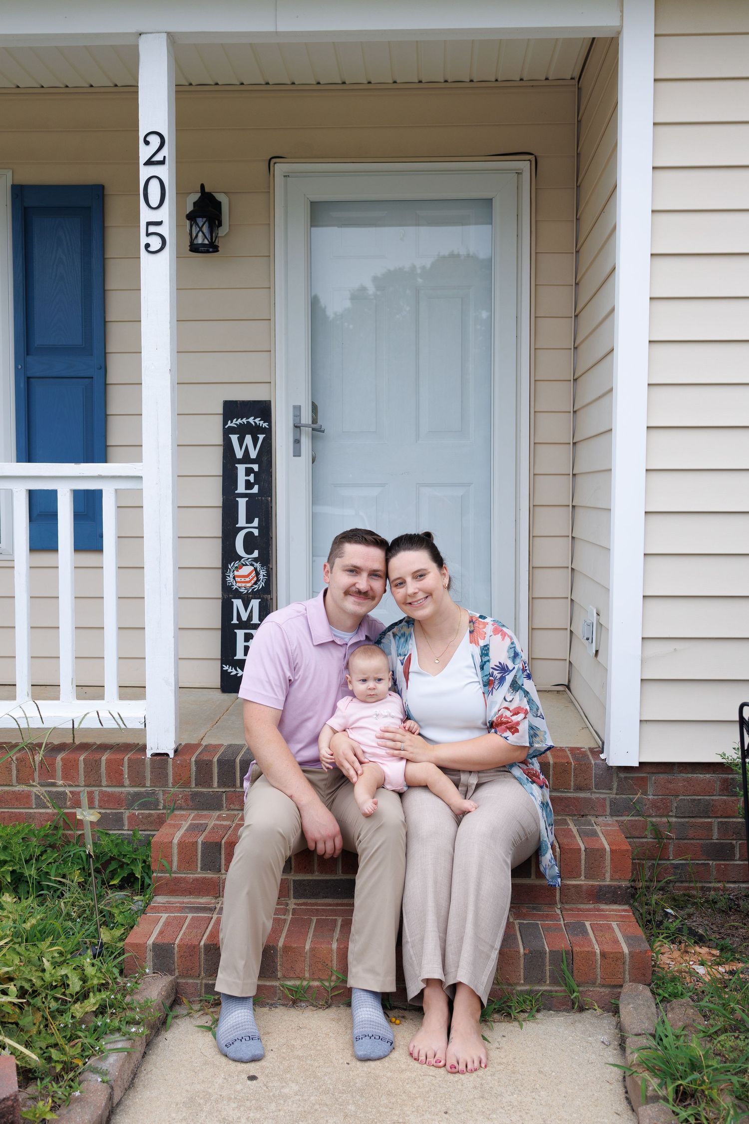 Smiling couple sits with their baby on the brick front steps of a beige house with a welcome sign and house number 205.
