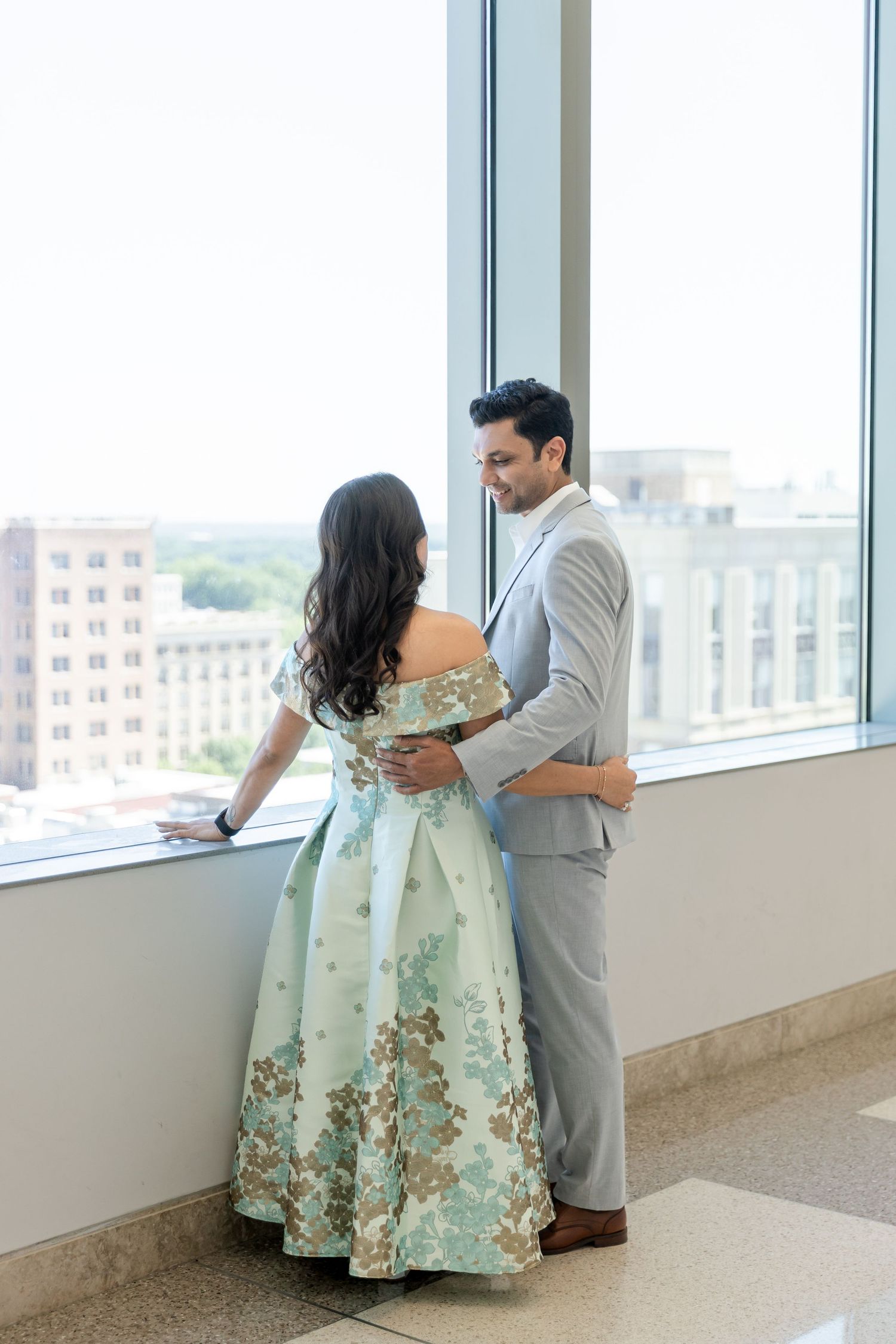 Couple in elegant attire embracing by large windows, woman in floral mint gown, man in gray suit, city view backdrop.Wake country courthouse