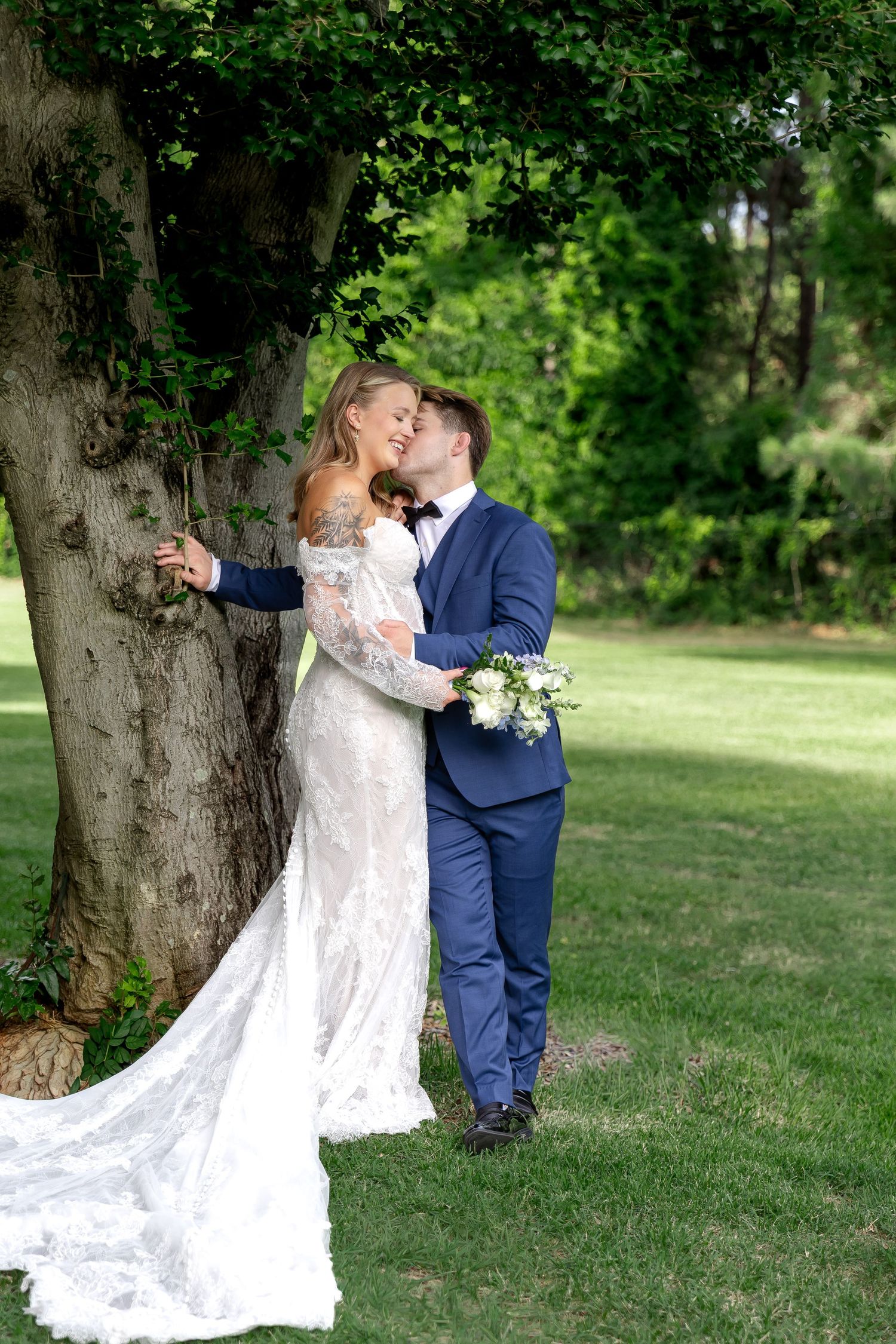 Bride and groom embracing under a large tree in a lush green garden, bride in lace gown holding white bouquet.