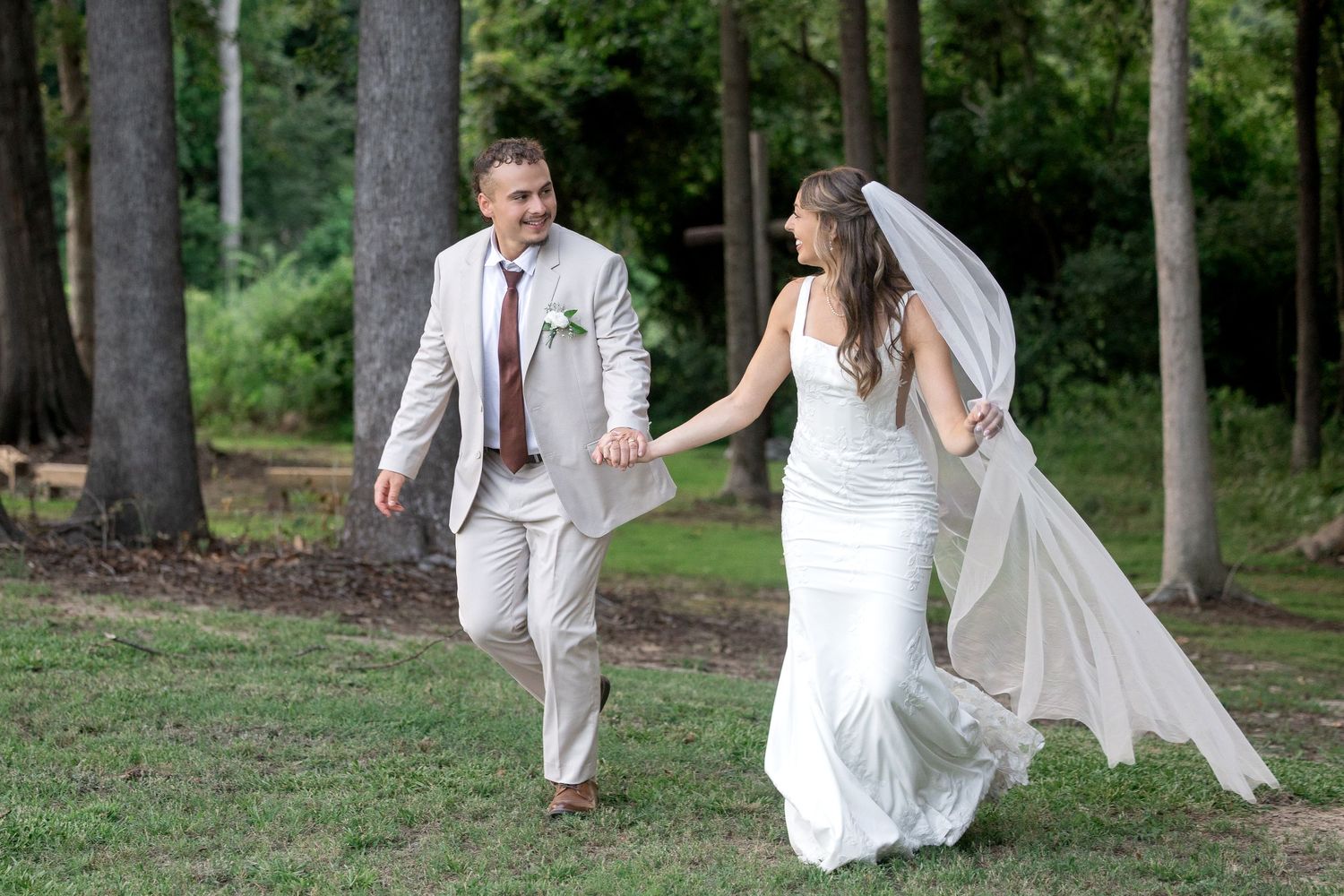 Bride in fitted lace gown and groom in tan suit hold hands walking through a wooded outdoor wedding venue.