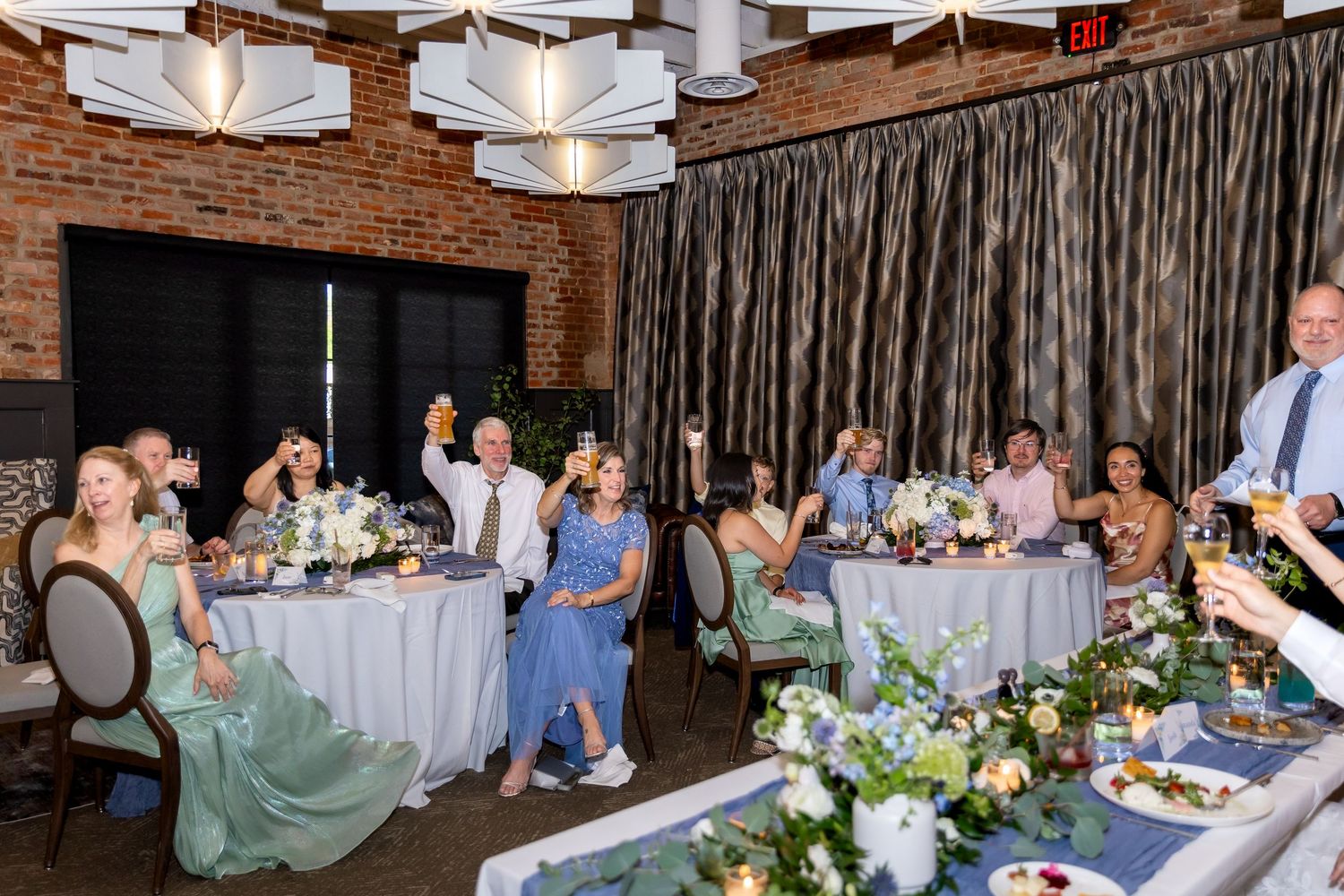 Wedding guests in formal attire raise glasses for a toast at floral-decorated round tables in a brick-walled reception venue.