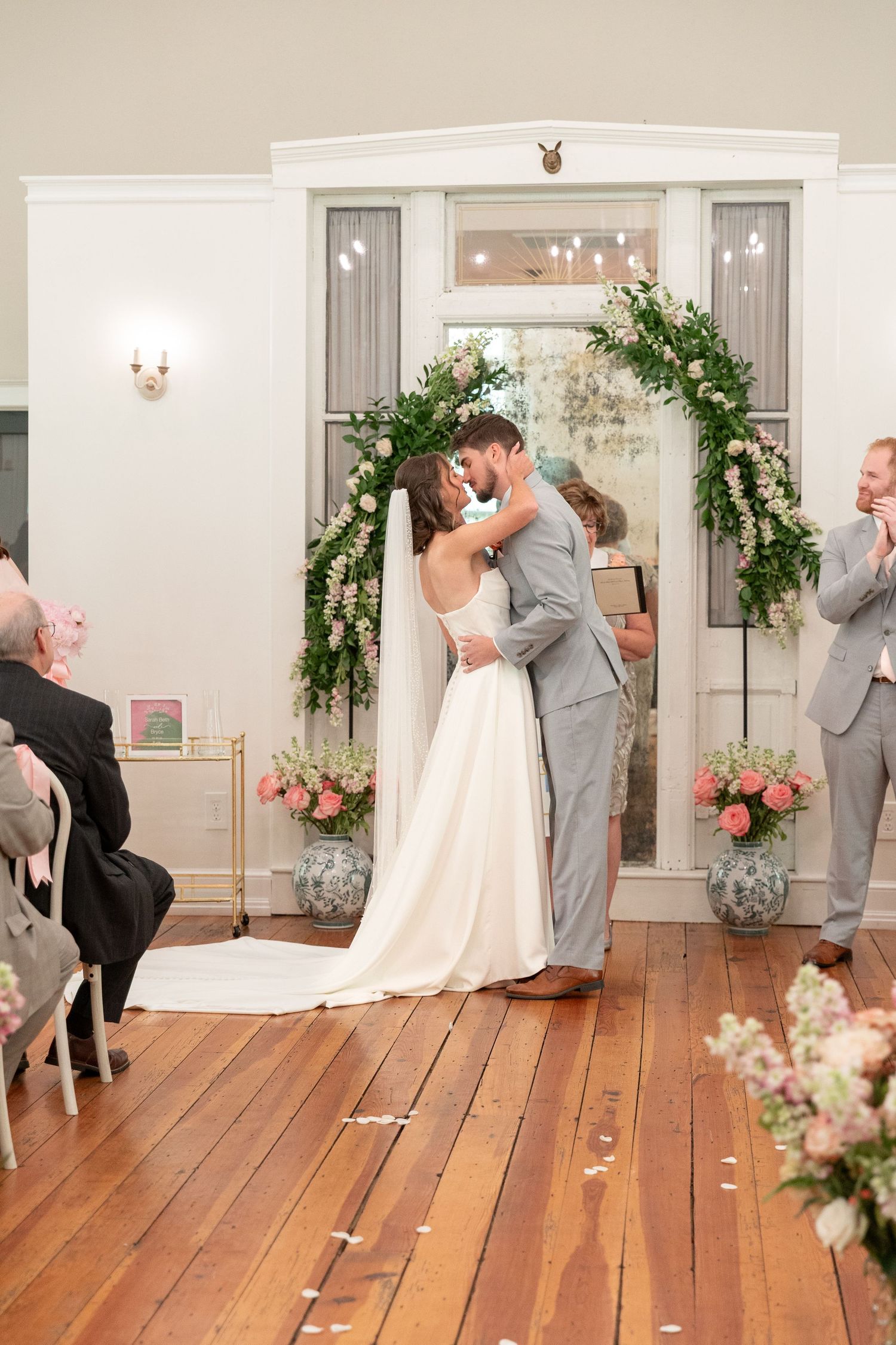 Bride and groom share a kiss during their wedding ceremony beneath a floral greenery arch, with guests and a smiling officiant looking on.