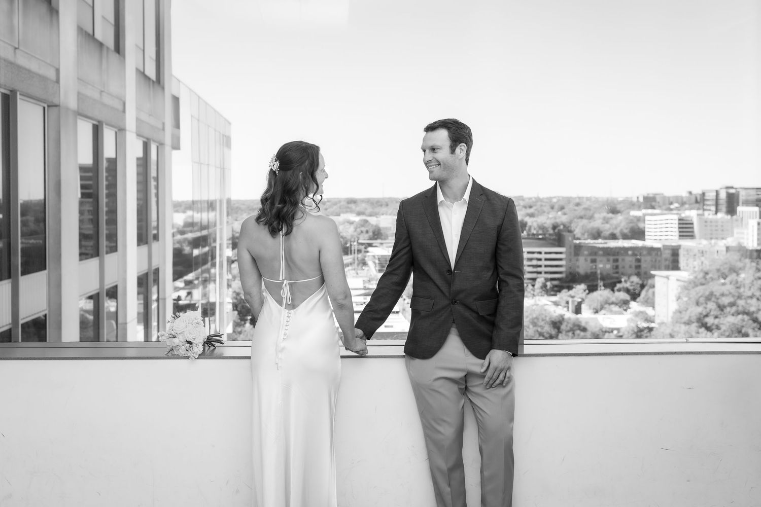Black and white photo of a bride and groom holding hands on a rooftop balcony with a city skyline backdrop.