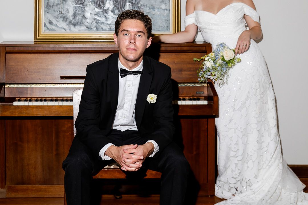Groom in black tuxedo sits at piano bench while bride in lace off-shoulder gown stands holding bouquet.