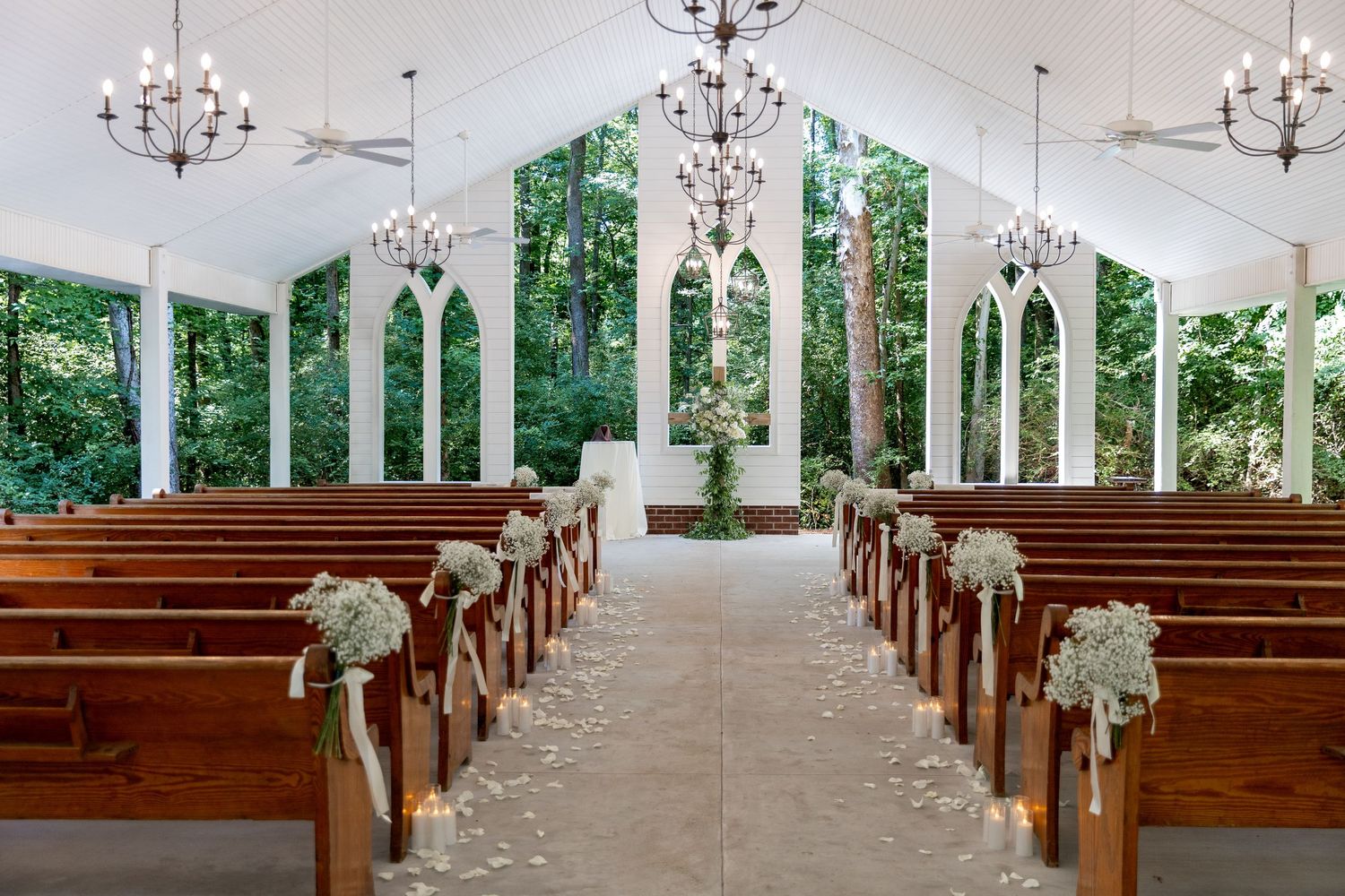 Stunning white chapel wedding aisle with wooden pews, floral arrangements, chandeliers, and arched windows.