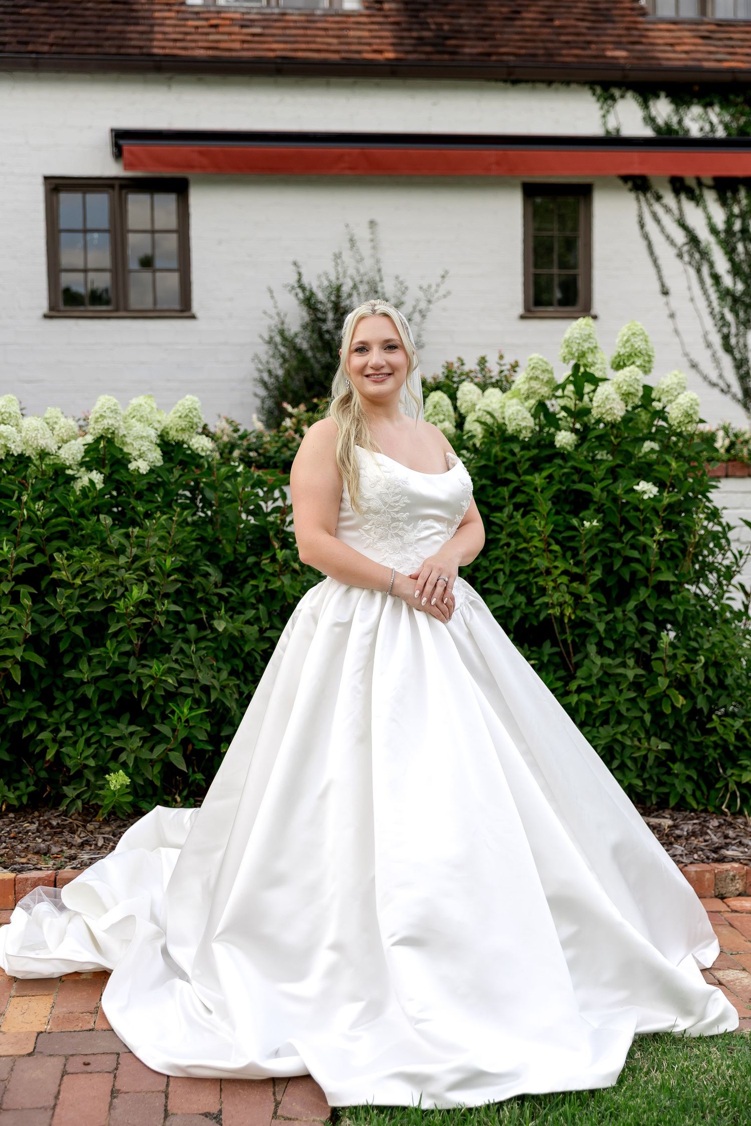 Smiling bride in a white ball gown wedding dress poses in a garden with white hydrangeas and a white brick building.