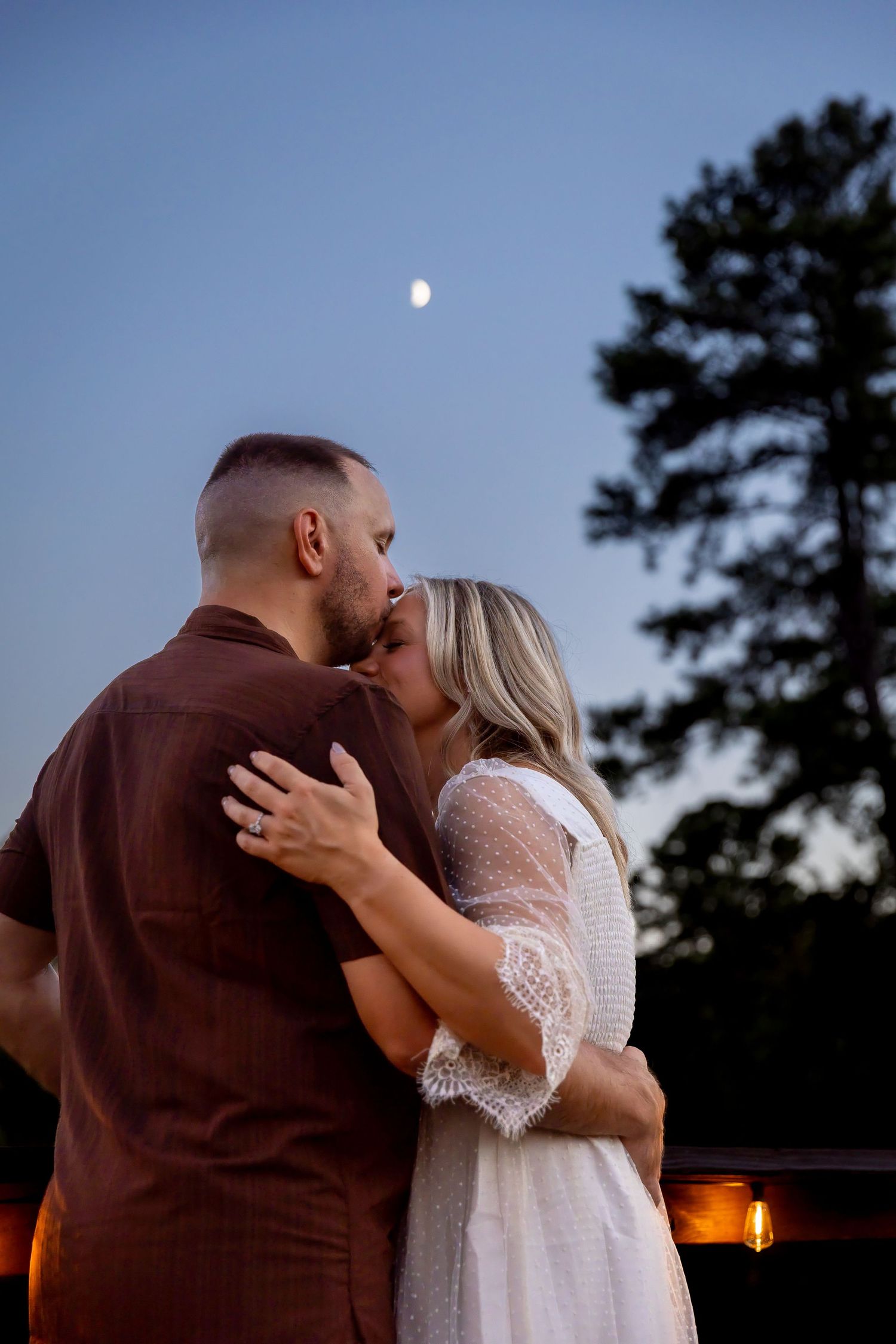 Couple embracing and kissing at dusk under a crescent moon, the woman in a white lace dress and the man in a brown shirt.