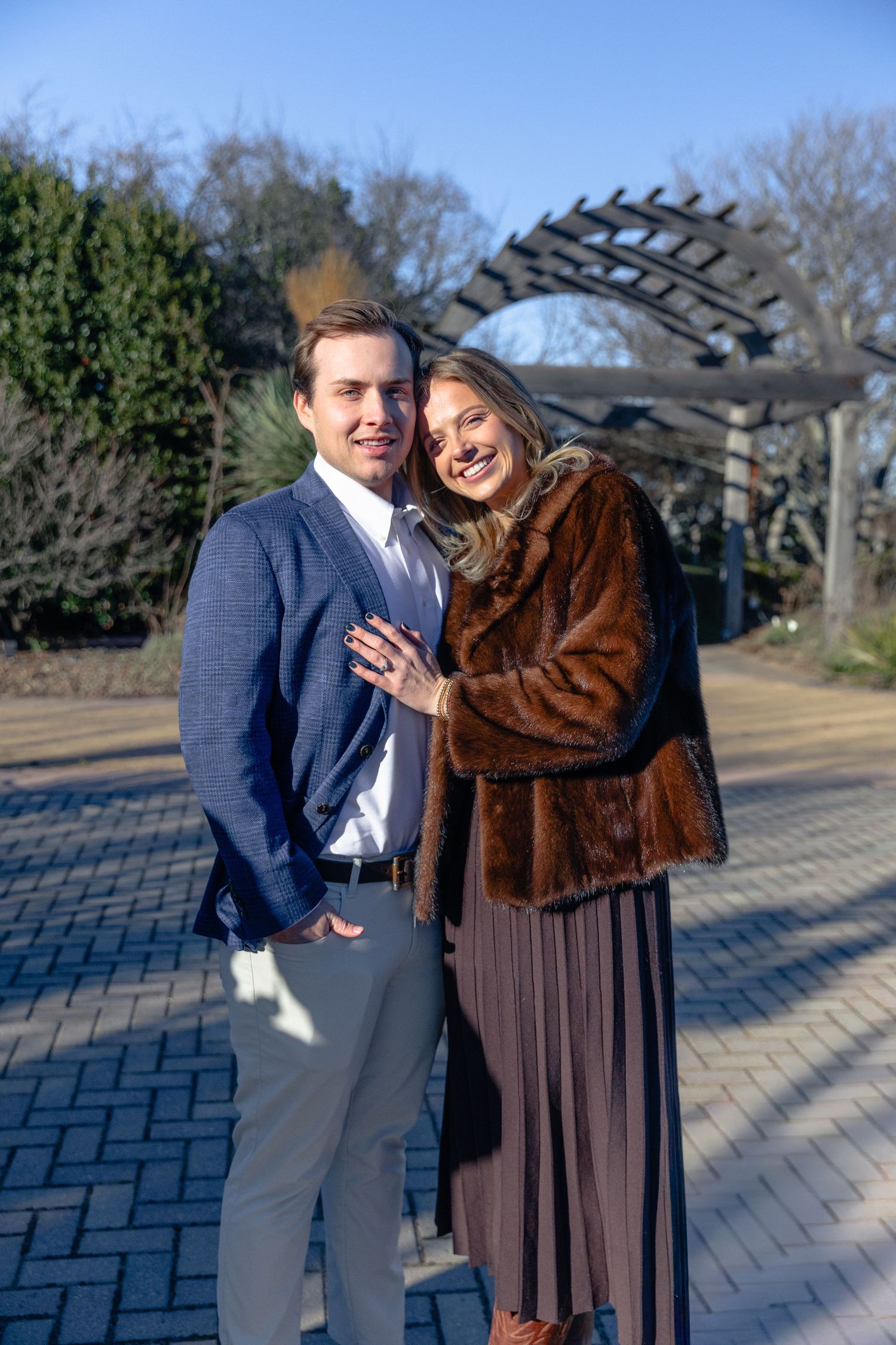 Smiling engaged couple posing outdoors in winter, woman in fur coat showing engagement ring.