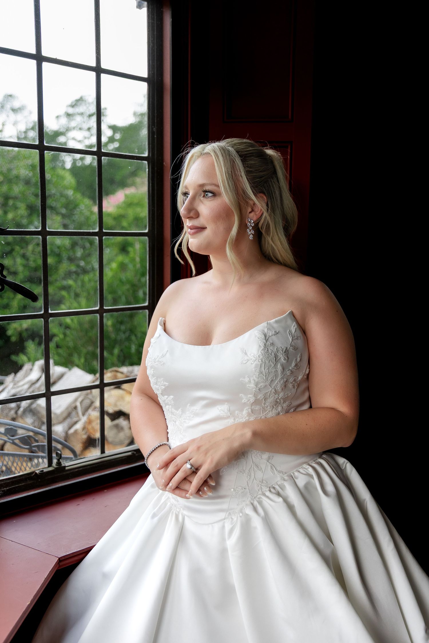 Bride in strapless white lace wedding gown gazing out a large window, wearing elegant drop earrings and engagement ring.