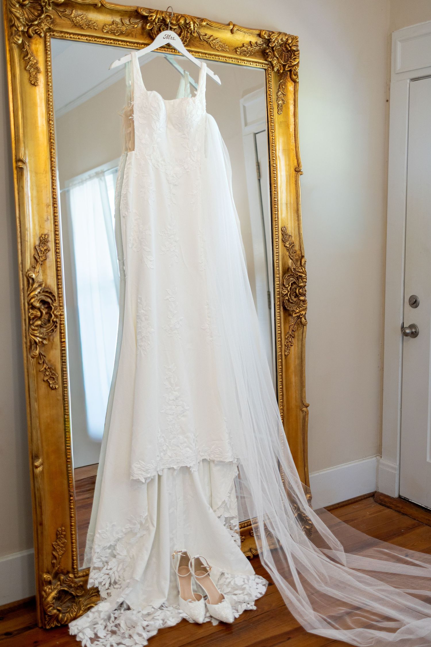 White lace wedding dress with long veil hanging on an ornate gold-framed floor mirror, with nude bridal heels resting below.