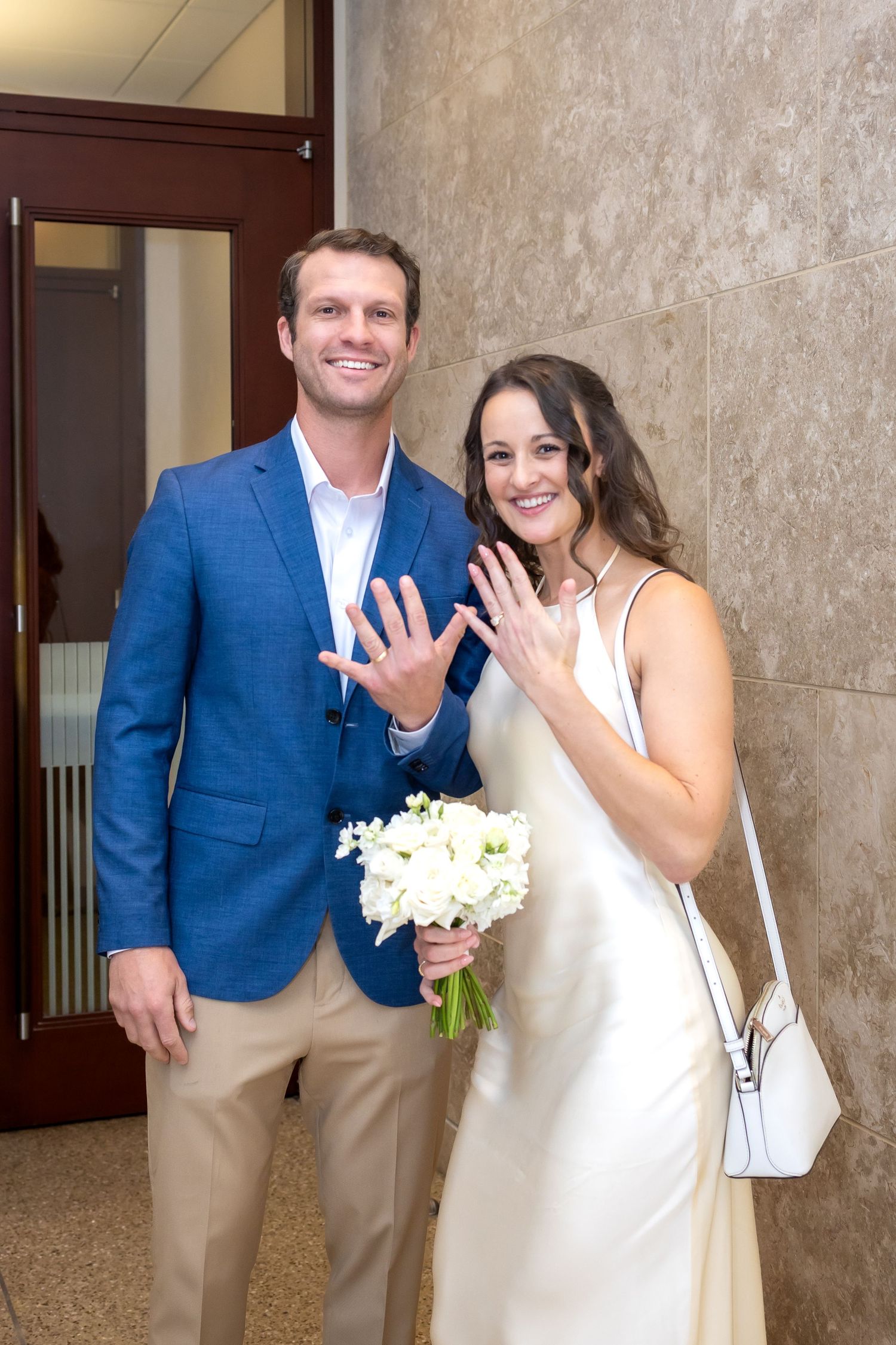 Newlywed couple smiling and showing off wedding rings, bride holding white bouquet in elegant satin dress, groom in blue blazer.