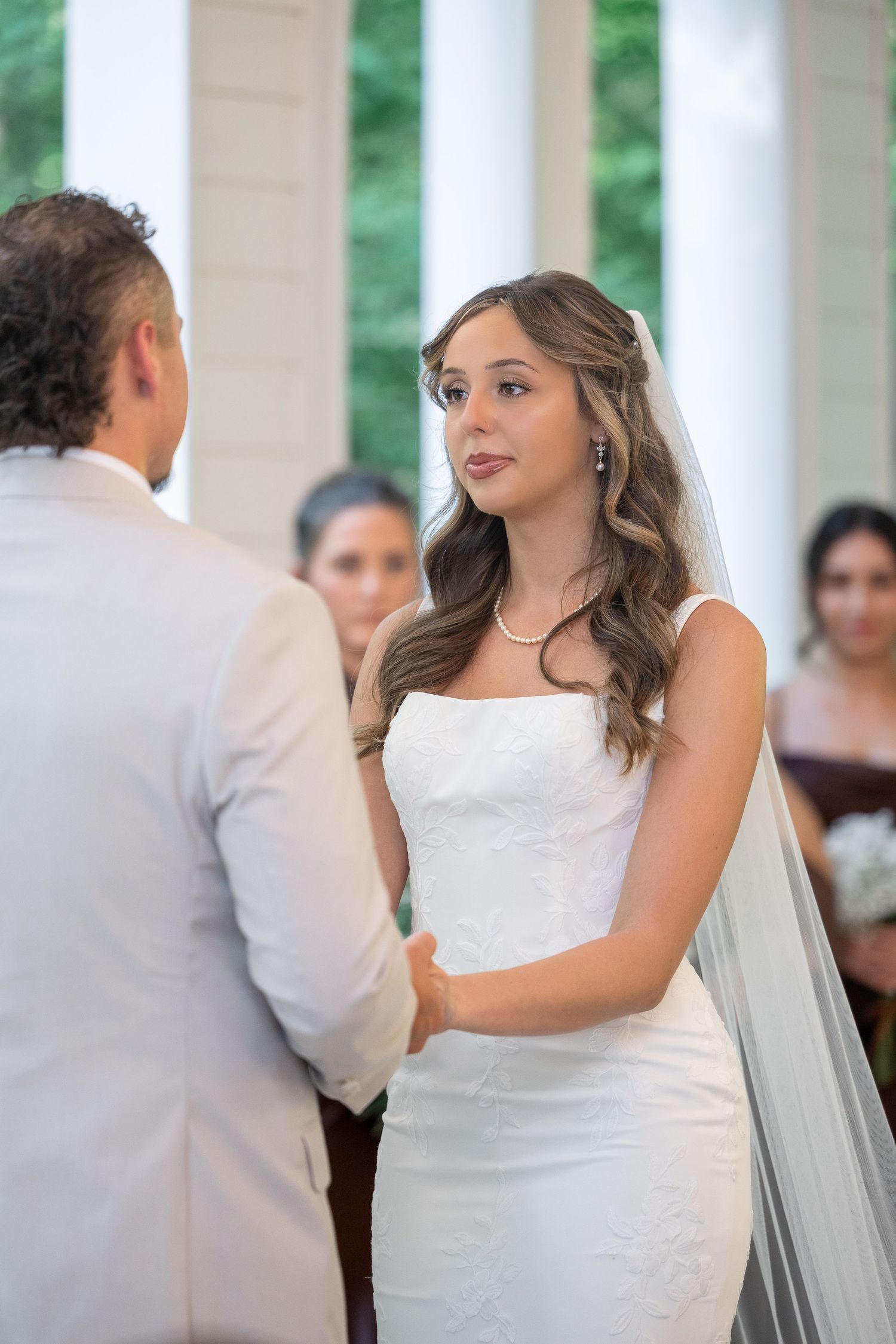 Bride in a strapless white gown and long veil holds hands with the groom during an outdoor wedding ceremony as guests look on.