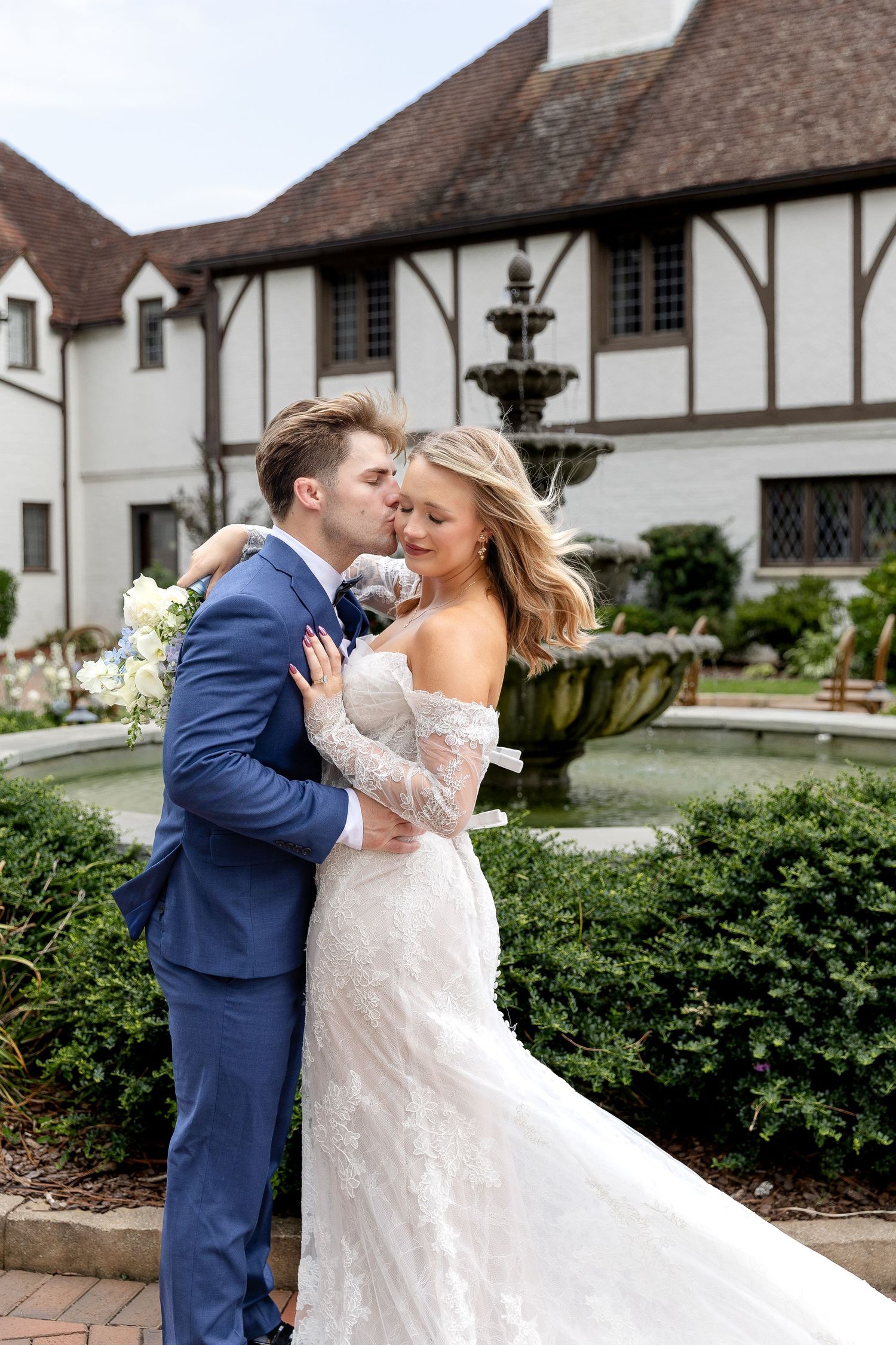 Bride and groom embrace by a stone fountain outside a Tudor-style venue, she holds white flowers in a lace gown.