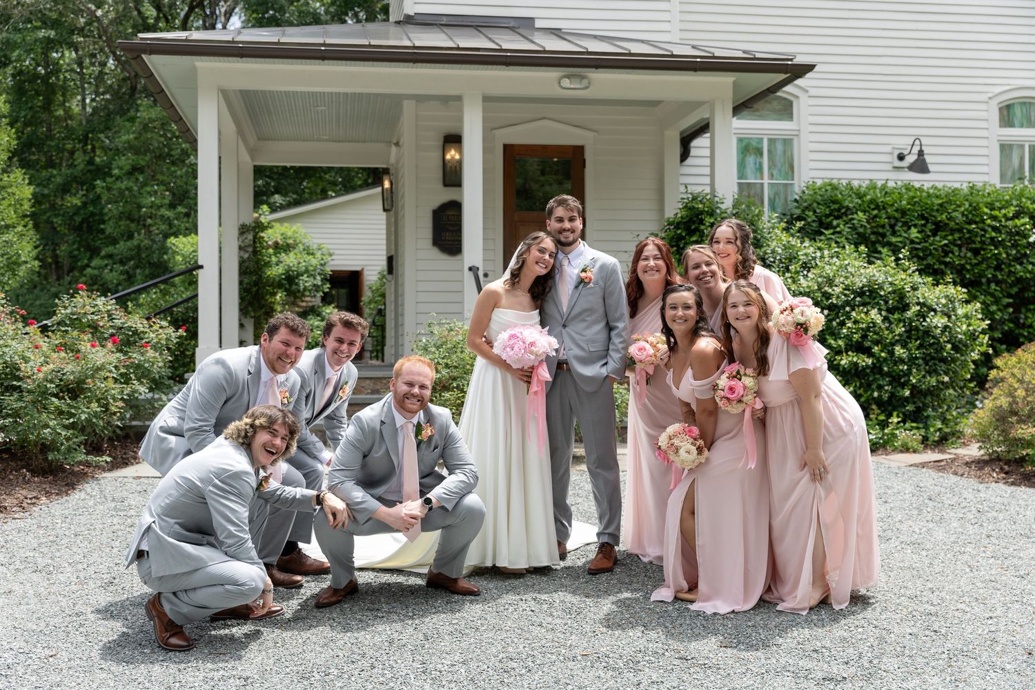 Bride and groom pose with bridesmaids in blush dresses and groomsmen in gray suits outside a white house on a gravel driveway.