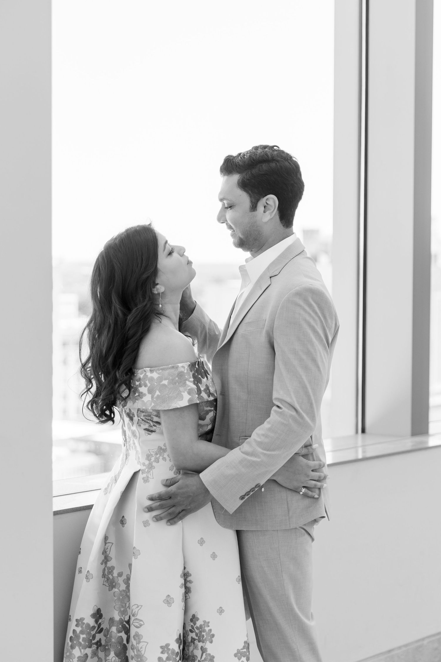 Black and white photo of a couple sharing a romantic moment by a large window, man in suit holding woman in floral dress.Wake country courth