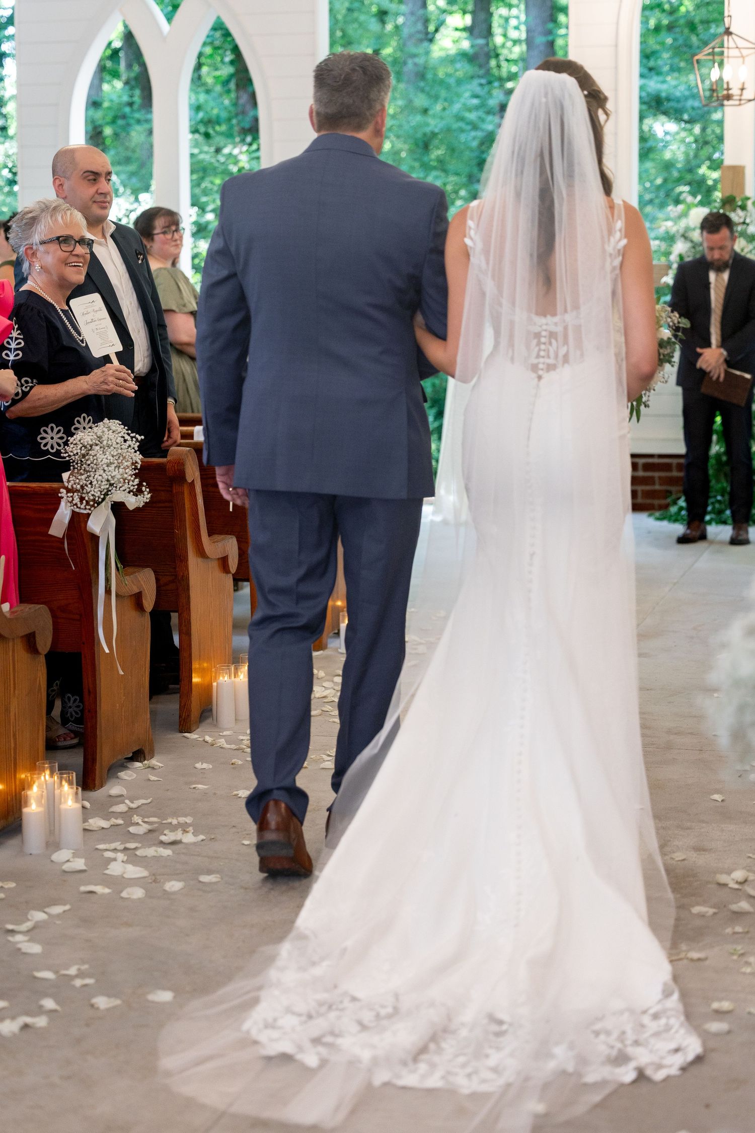 A bride in a lace mermaid gown with a long veil walks down a petal-strewn church aisle on her father's arm as guests watch.