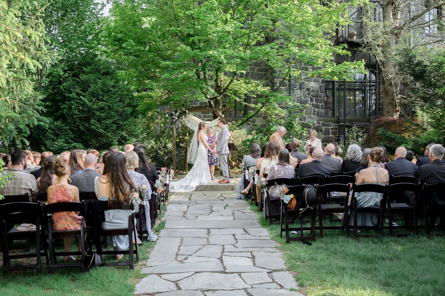 Outdoor garden wedding ceremony with guests seated in black chairs on stone pathway surrounded by lush green trees.