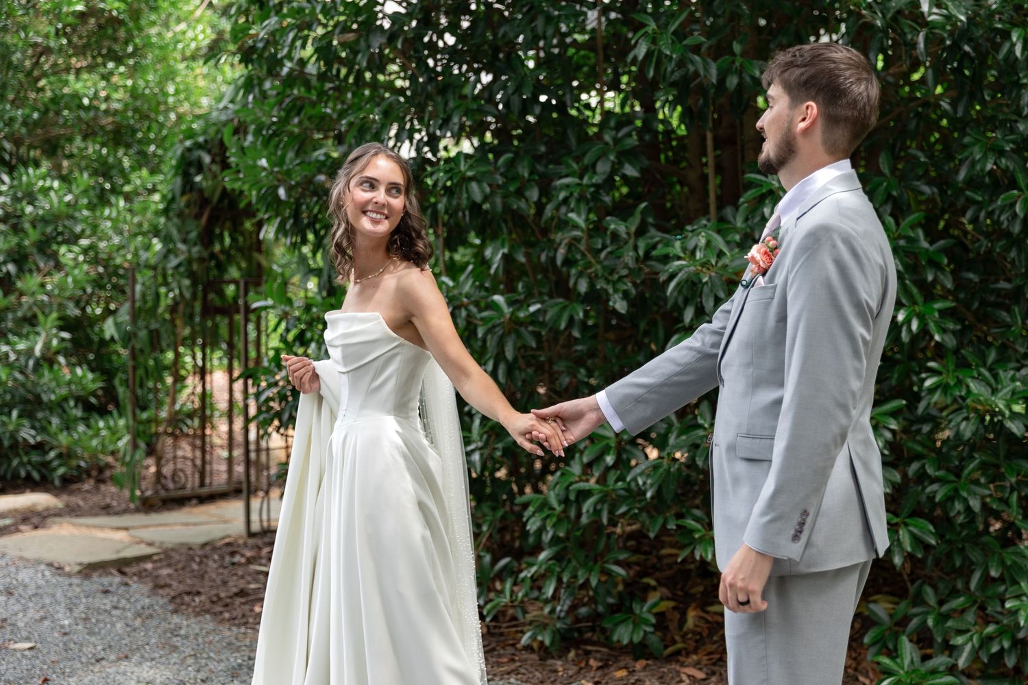 Smiling bride in a flowing white dress holds hands with a groom in a gray suit during a first look surrounded by lush green foliage.