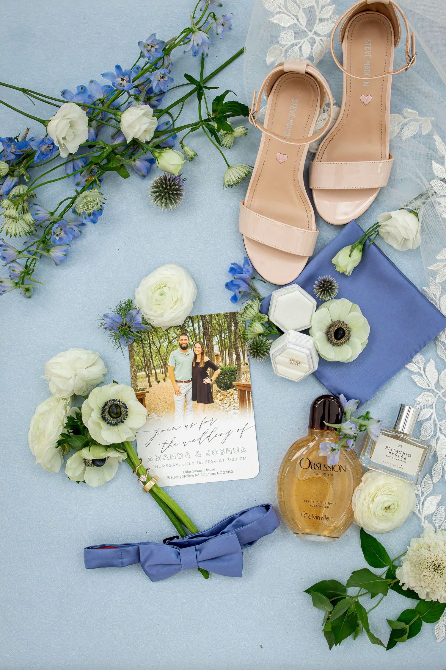 Wedding flat lay with blush heels, blue bow tie, perfume, ring boxes, invitation and white and blue flowers.