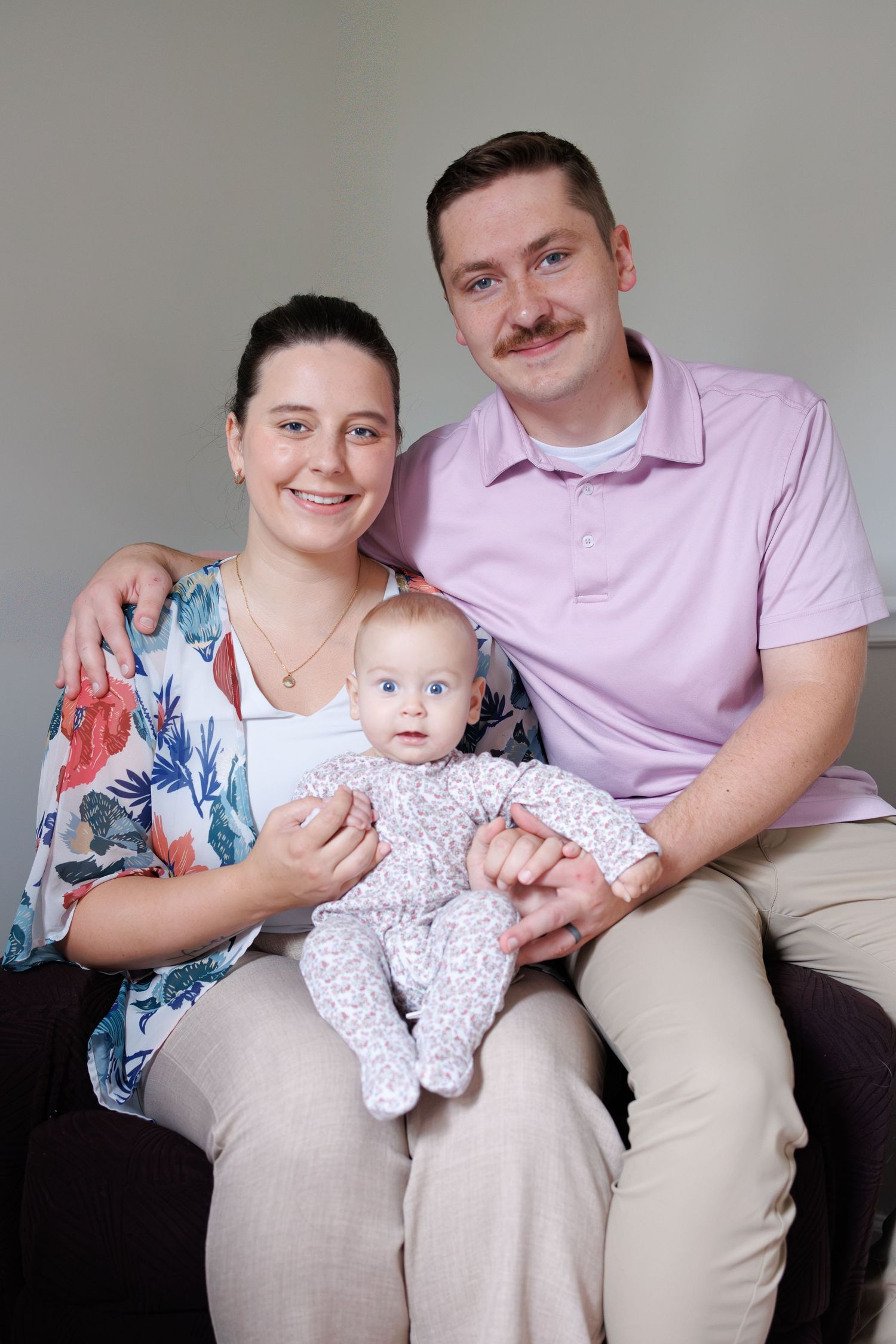 Smiling woman and man with a mustache sit together on a couch holding their baby, who wears a floral onesie, in a bright family portrait.