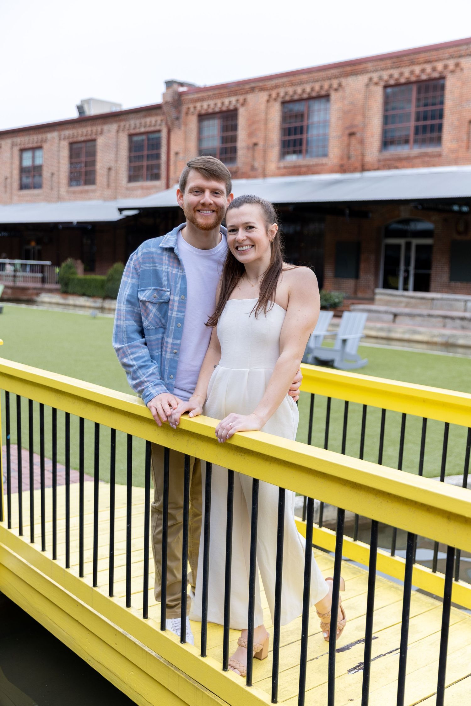 Happy couple leaning on yellow railing of a bridge outside a rustic brick building.