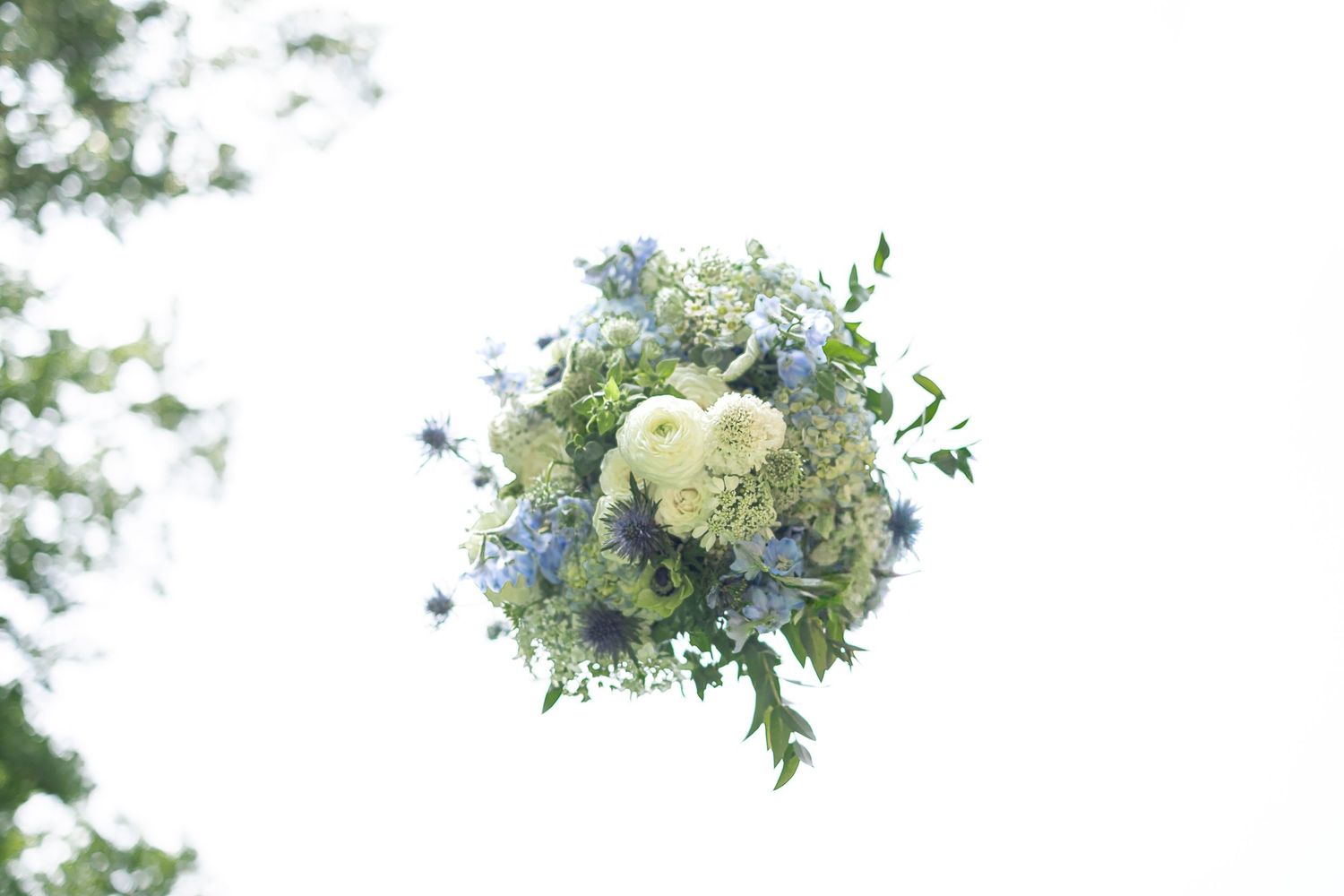 Delicate blue and white floral wedding bouquet with greenery floating against a bright white sky.