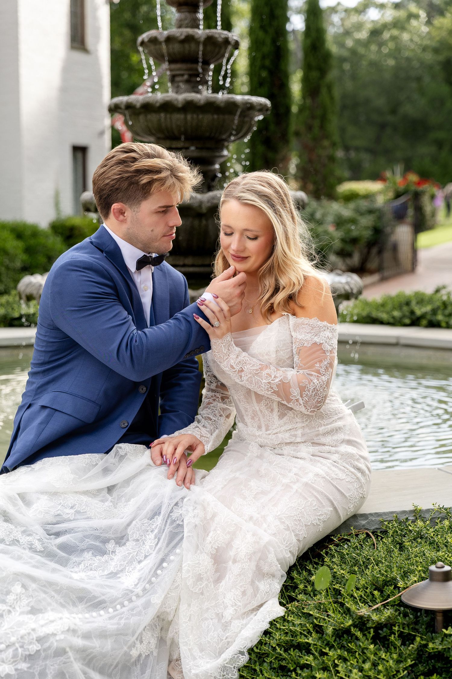 A groom in a blue suit tenderly holds his bride's hand by a stone fountain outdoors.