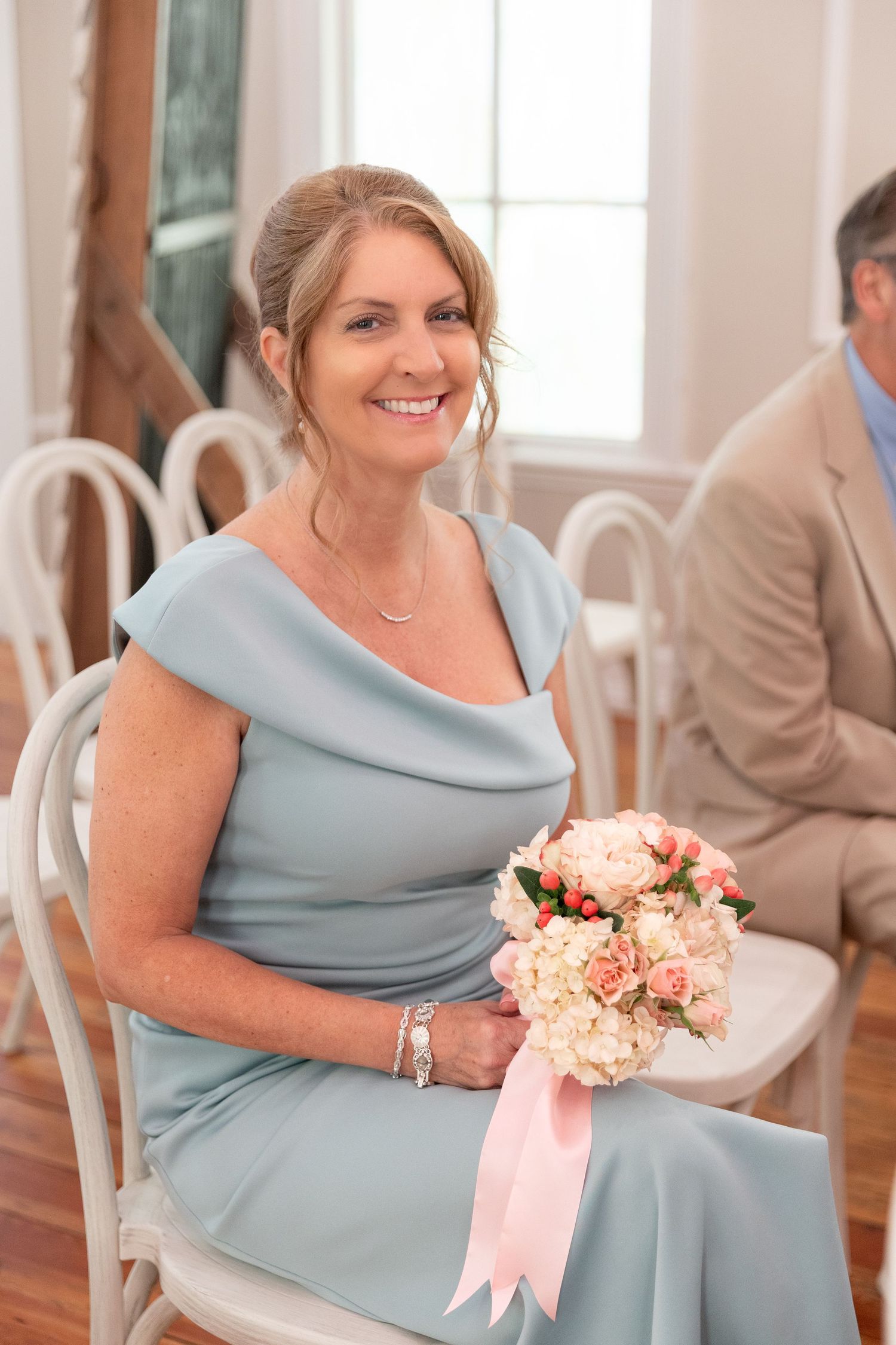 Smiling woman in a dusty blue off-shoulder dress holds a pink and white rose bouquet while seated on a white chair at a wedding.