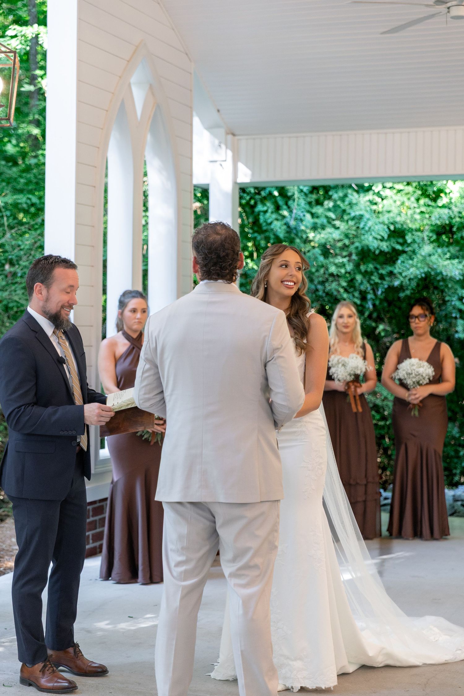 A smiling bride in a white gown and veil faces her groom during an outdoor wedding ceremony as an officiant and bridesmaids look on.