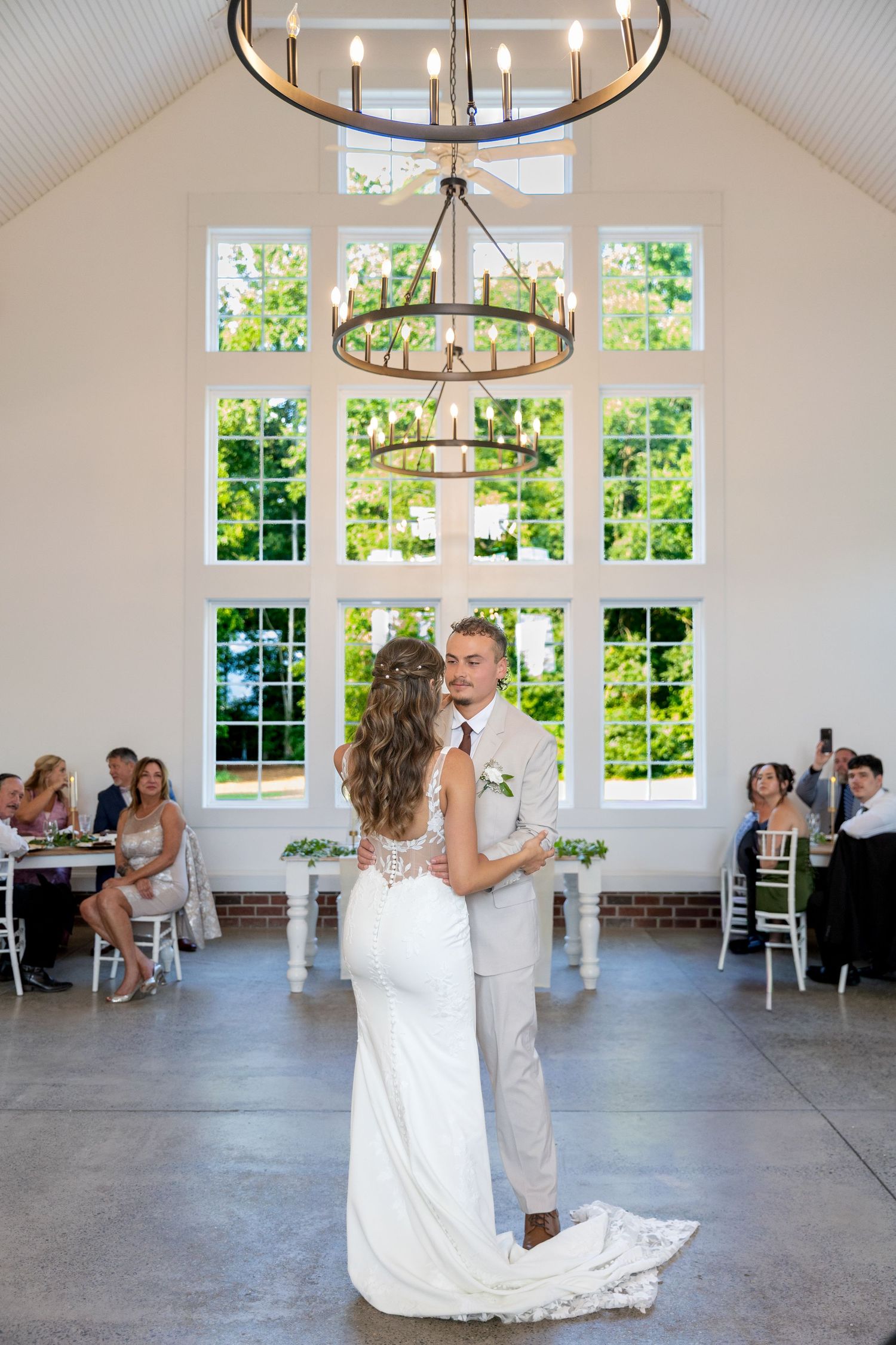 Bride and groom sharing first dance under chandelier in bright white venue with large windows.
