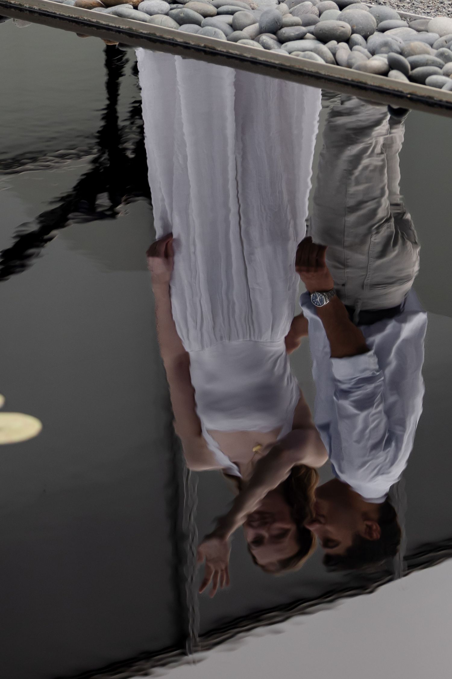 Couple in white wedding attire reflected upside down in still water near pebble shore.